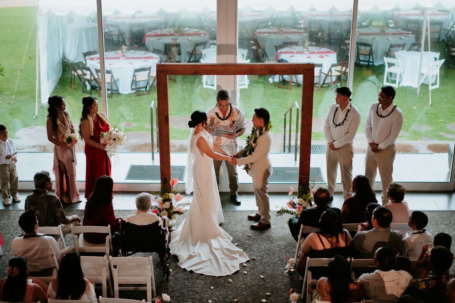 Wedding ceremony indoors with couple exchanging vows at altar, bridesmaids in red, groomsmen in tan, guests seated.