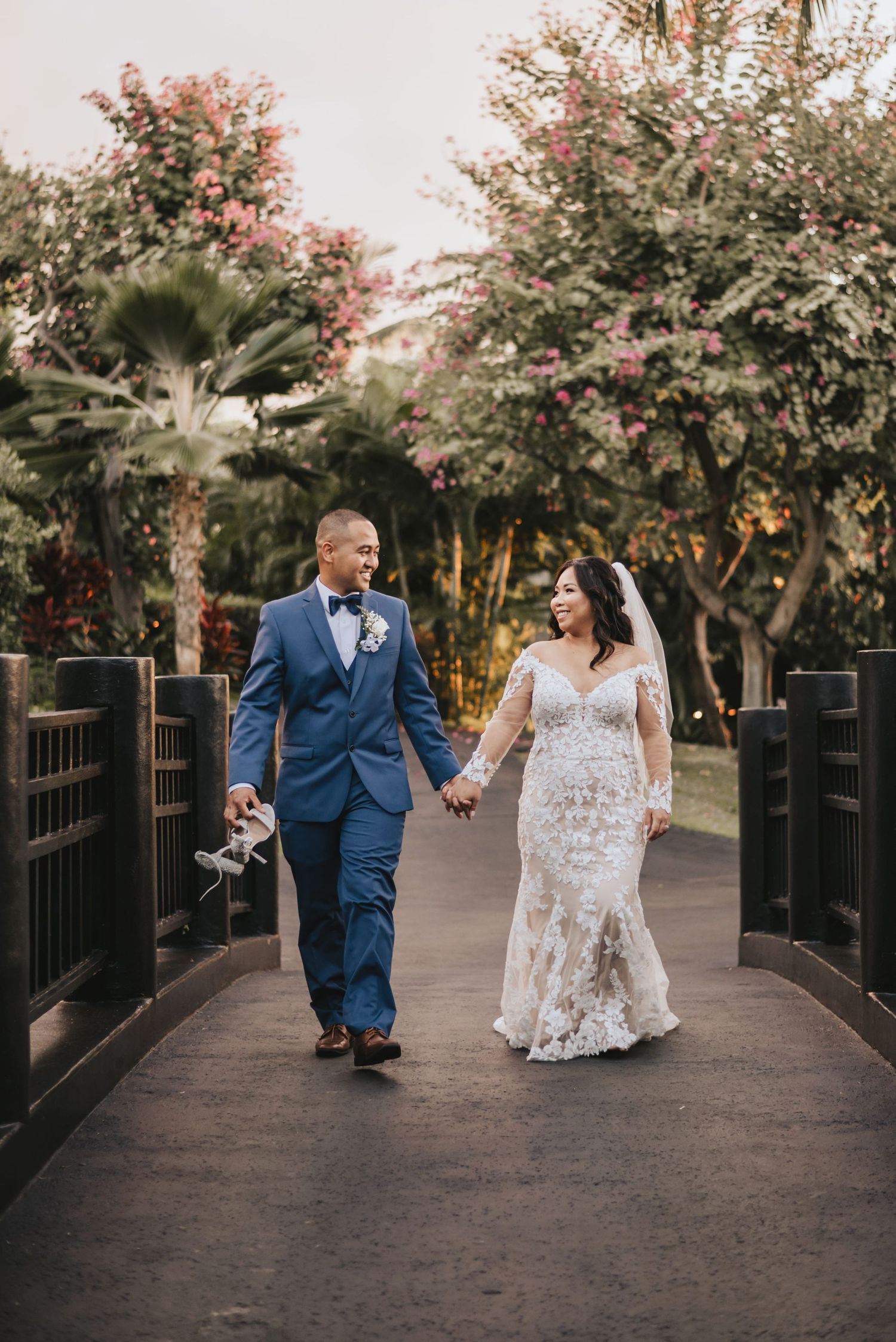 Bride and groom walk hand-in-hand on a bridge surrounded by lush tropical trees and pink flowers.