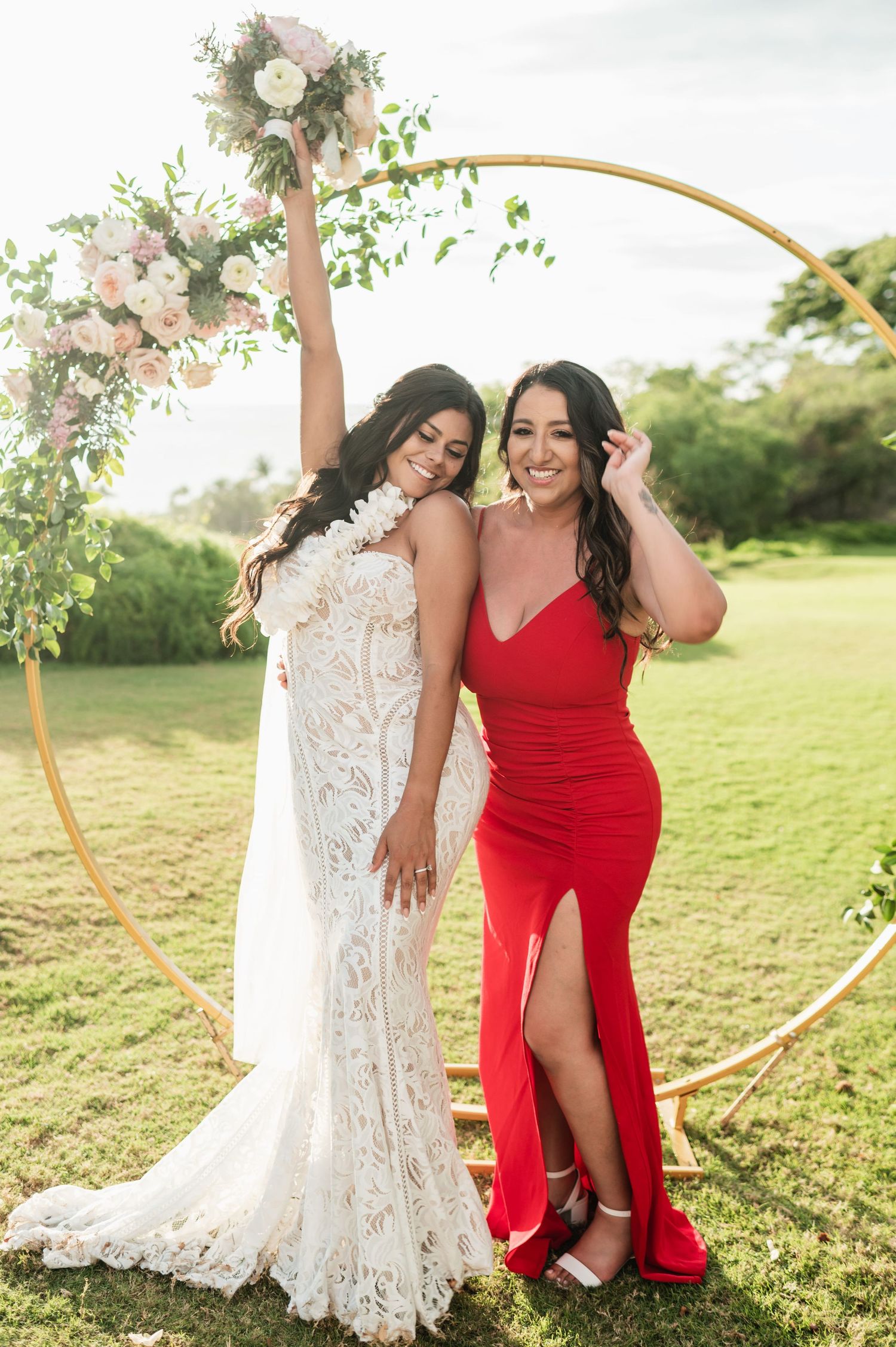 Bride in white lace gown and bridesmaid in red dress pose under floral hoop arch at outdoor wedding.