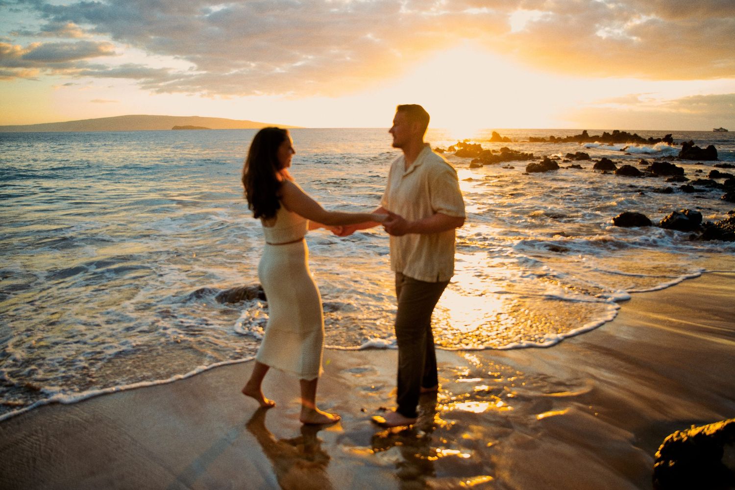 Romantic couple holding hands on beach at golden sunset with waves at their feet.