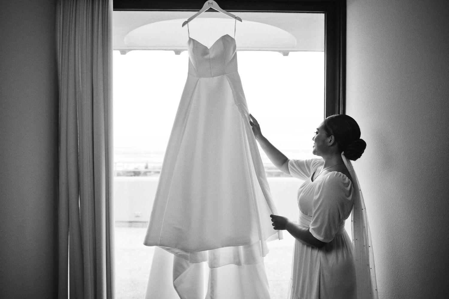 Bride in white robe admiring her wedding gown hanging by a sunlit window in a black and white photo.
