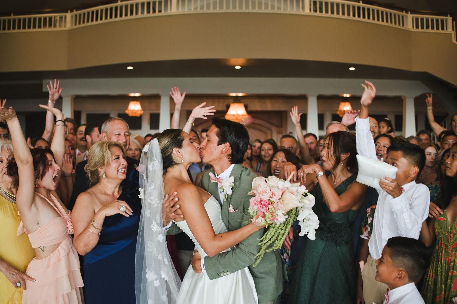 Bride and groom share a kiss surrounded by cheering wedding guests in an elegant venue.