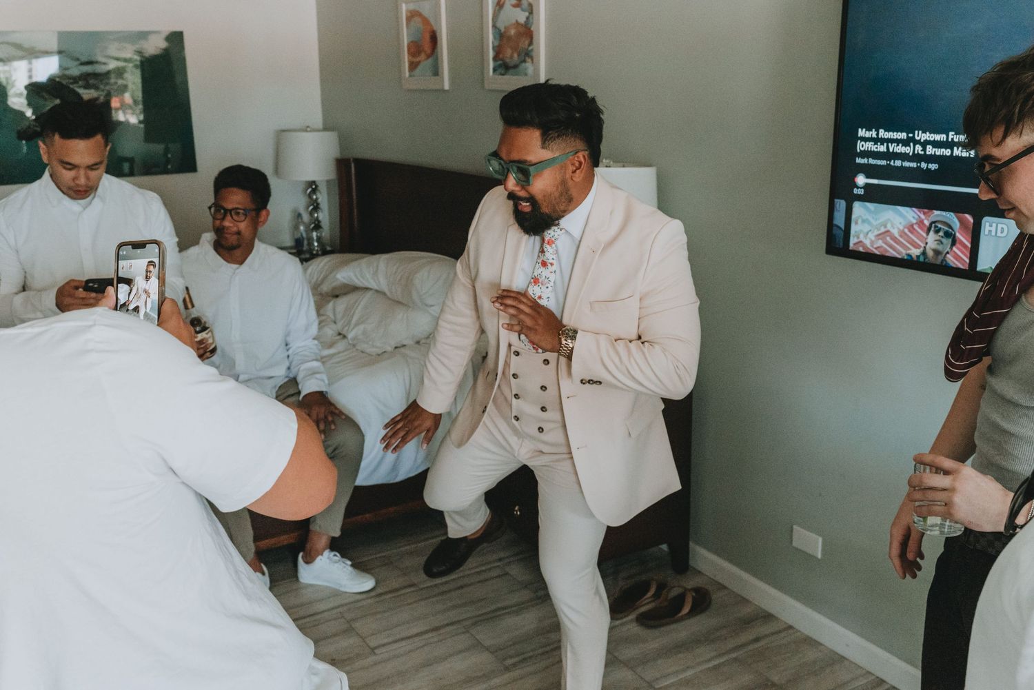 Groom in white suit dancing with groomsmen in hotel room before wedding ceremony.