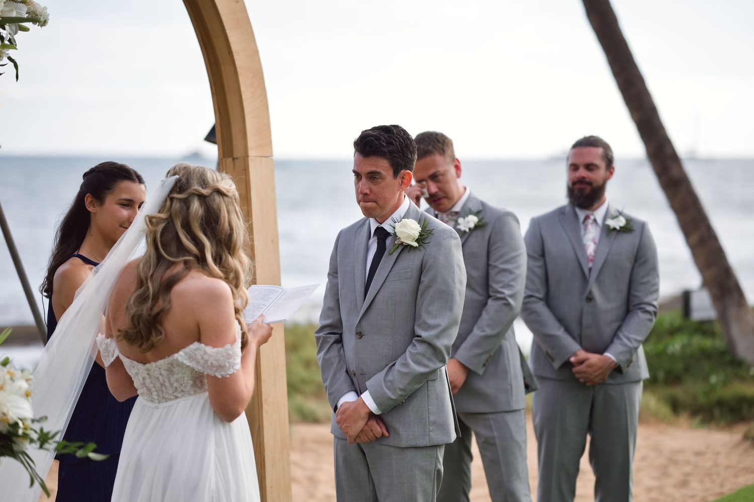 Groom in gray suit exchanges vows with bride in white lace dress at outdoor beach wedding ceremony with groomsmen.