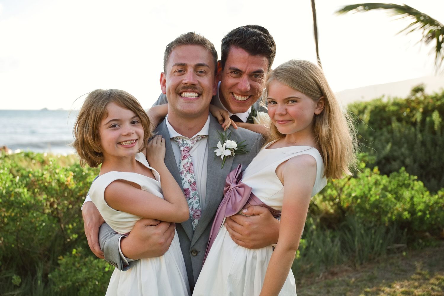 Groom and bestman in suits smile with two flower girls in white dresses at an outdoor beach wedding ceremony.