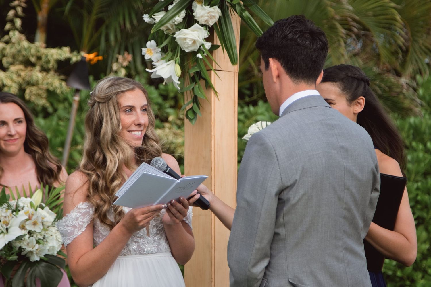 Bride smiling during outdoor wedding ceremony, holding vows booklet, with floral arch backdrop.