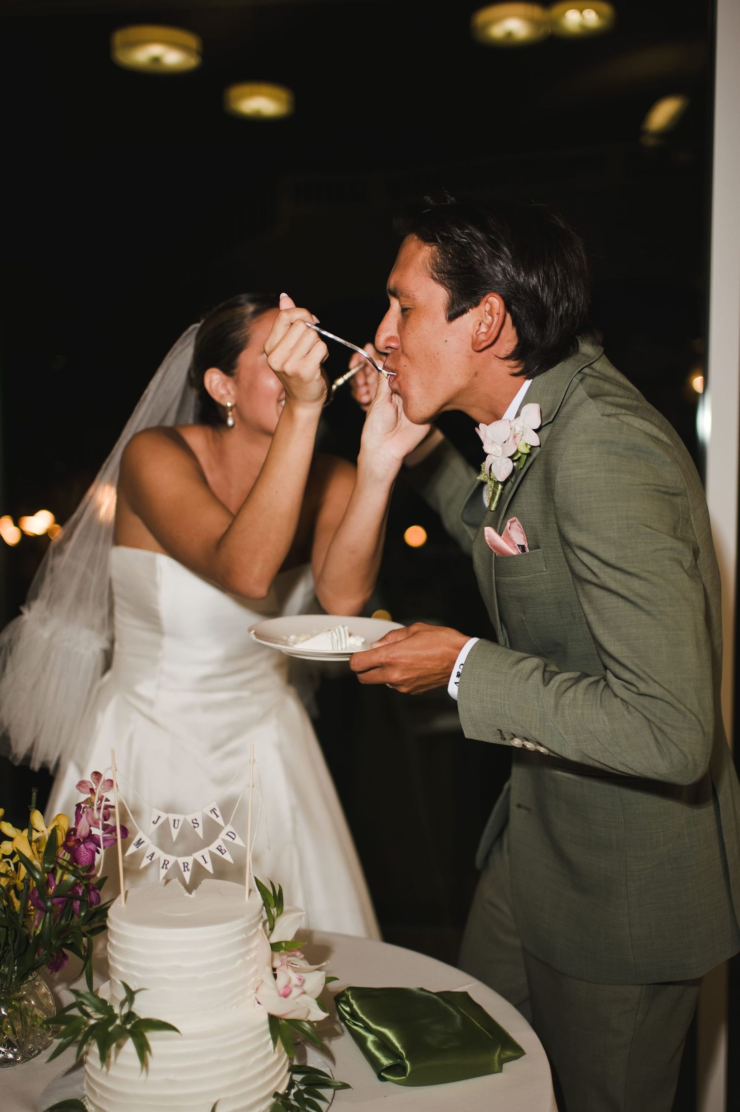 Bride and groom share first bite of white wedding cake, groom in green suit, bride in white gown with veil.