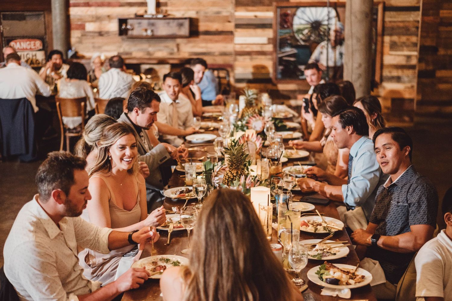 Wedding reception dinner with guests seated at long tables in rustic wooden venue.
