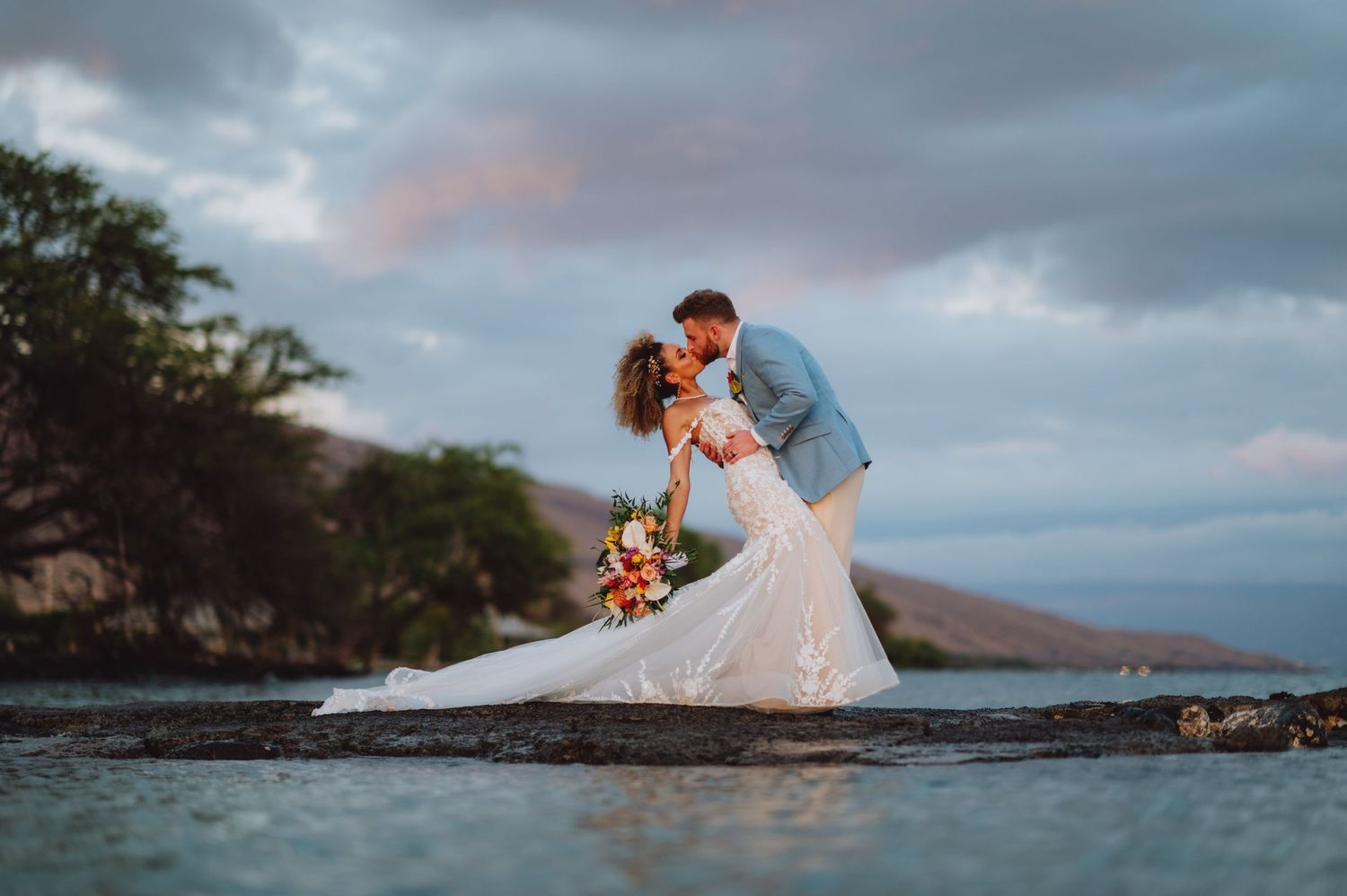 Dramatic cloudy sky frames wedding couple on Hawaiian beach at sunset.