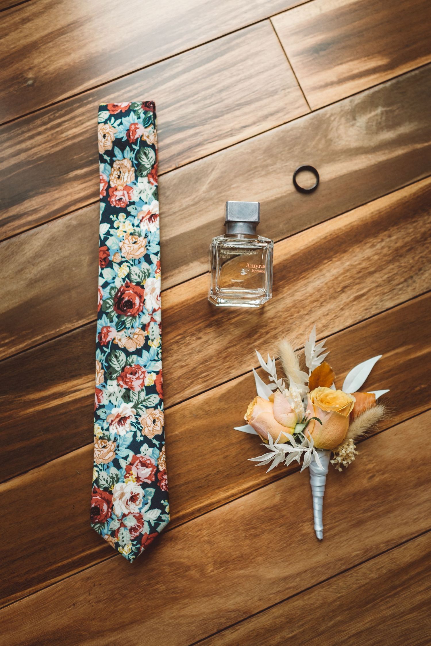 Wedding accessories including floral tie, perfume bottle and boutonniere on wooden floor.