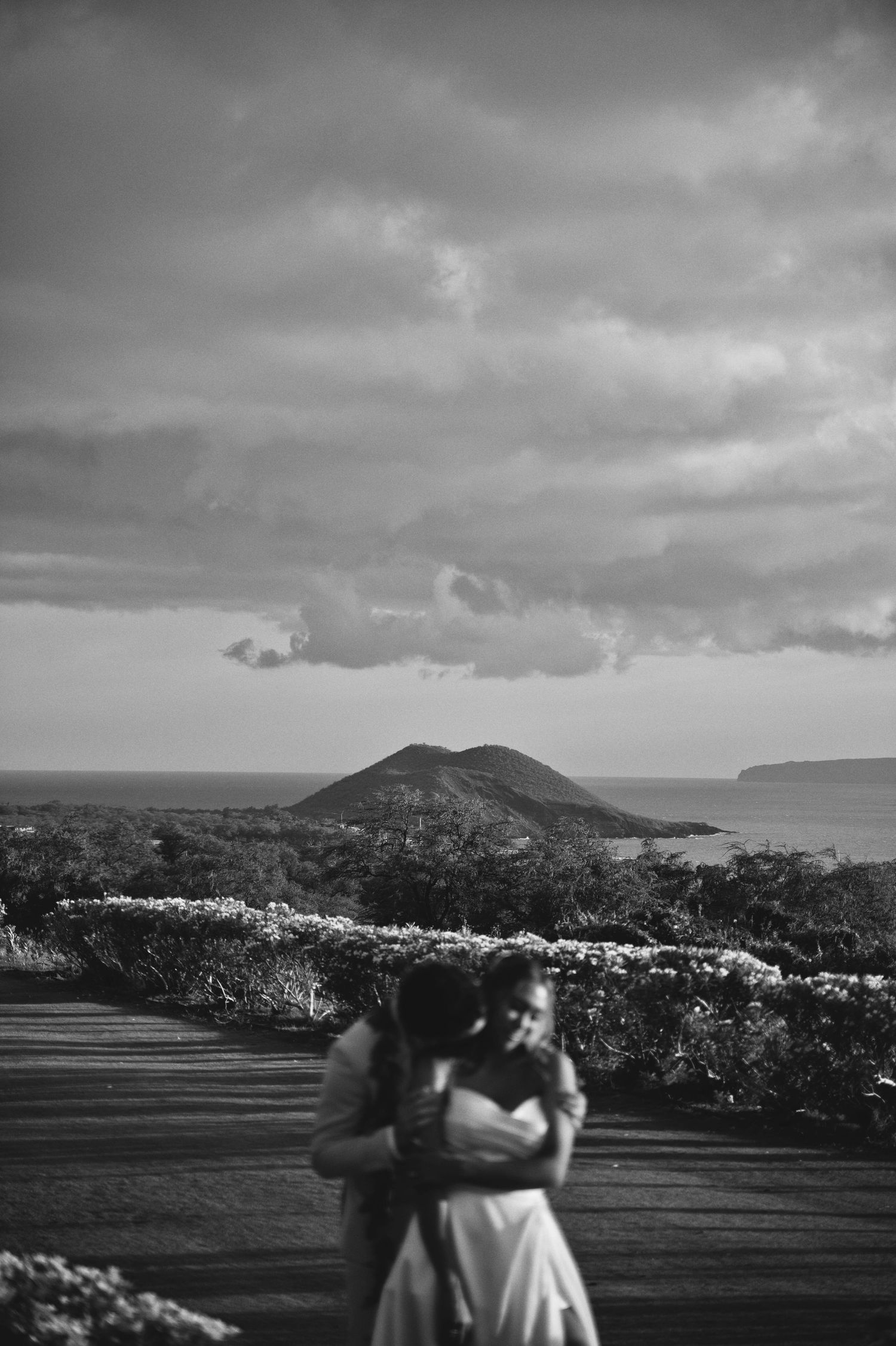 Black and white romantic photo of couple embracing on deck with volcanic island and ocean backdrop.