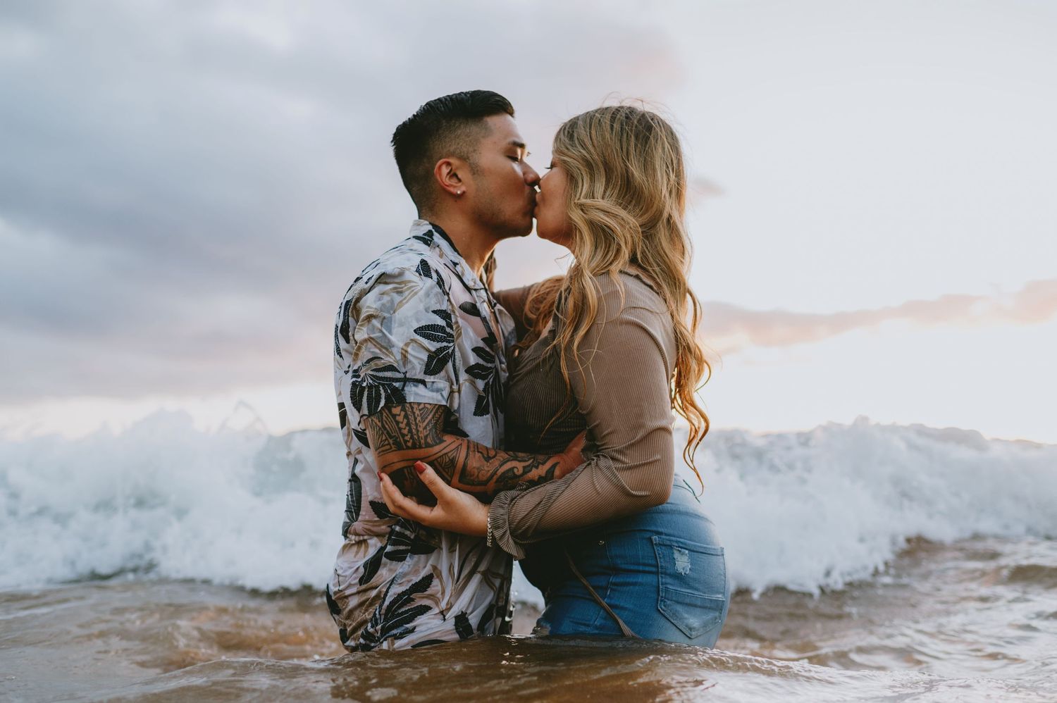 Couple sharing a passionate kiss while sitting in ocean waves on a beach.