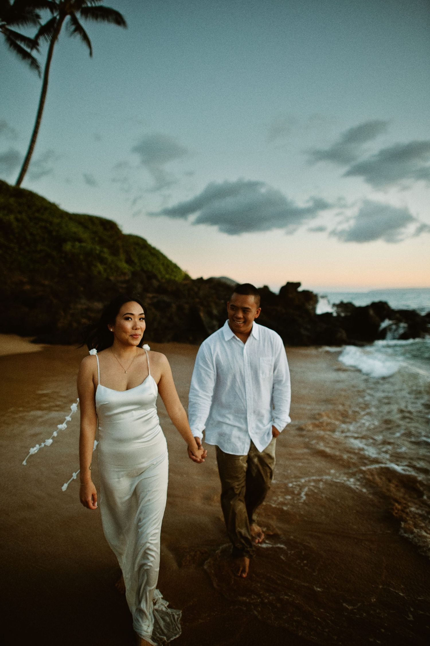 Couple holding hands walking along a sandy beach at sunset with palm trees.