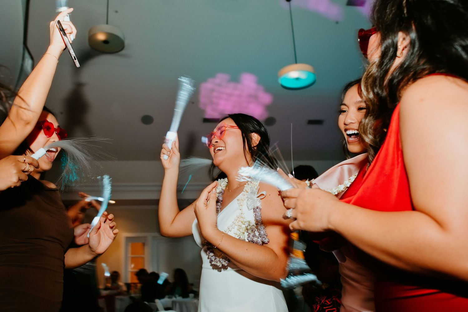 Bride dancing joyfully with friends holding glow sticks at a lively wedding reception party.