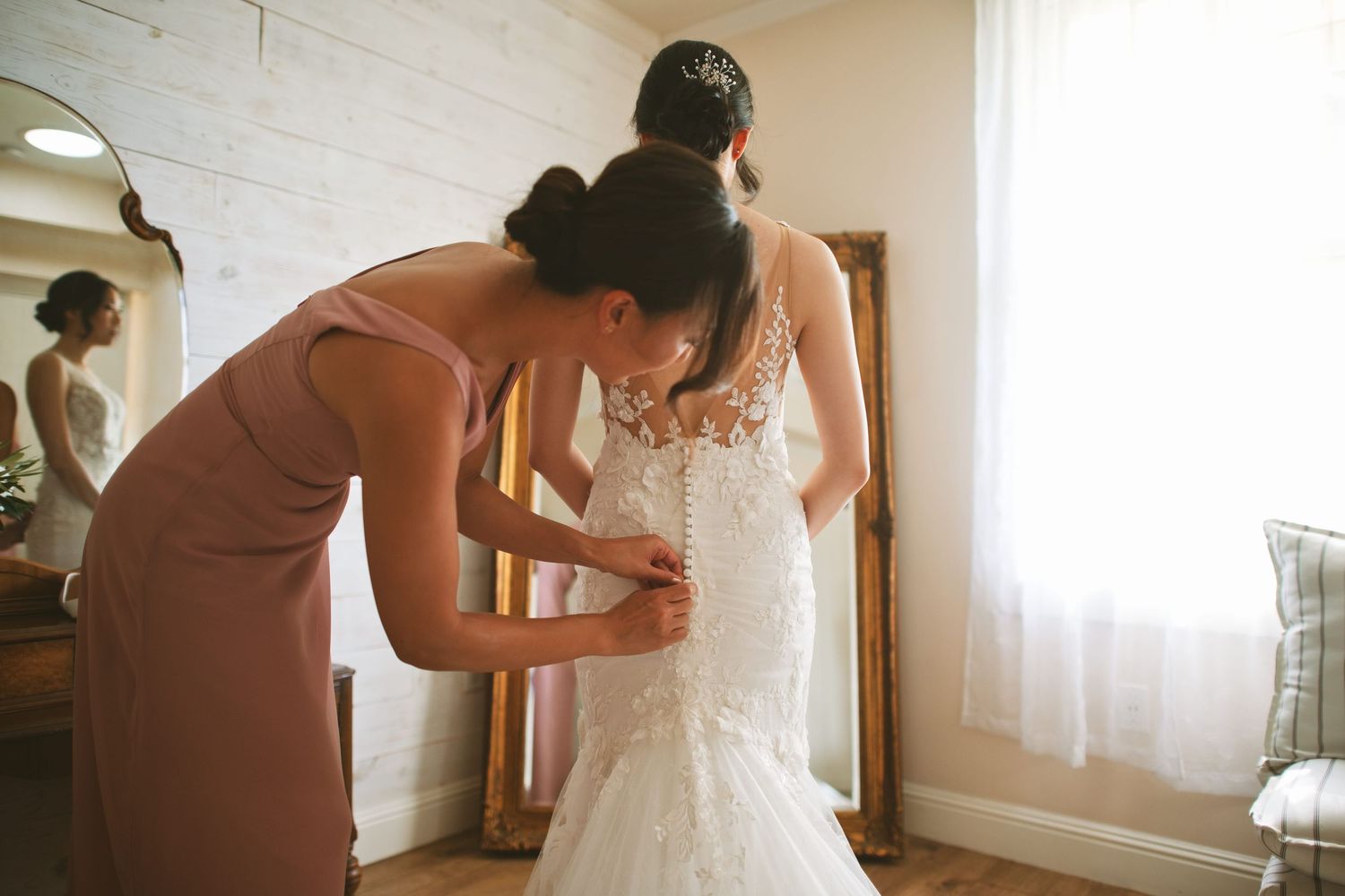 Bridesmaid in dusty rose dress helping bride fasten lace mermaid wedding gown in bright dressing room.