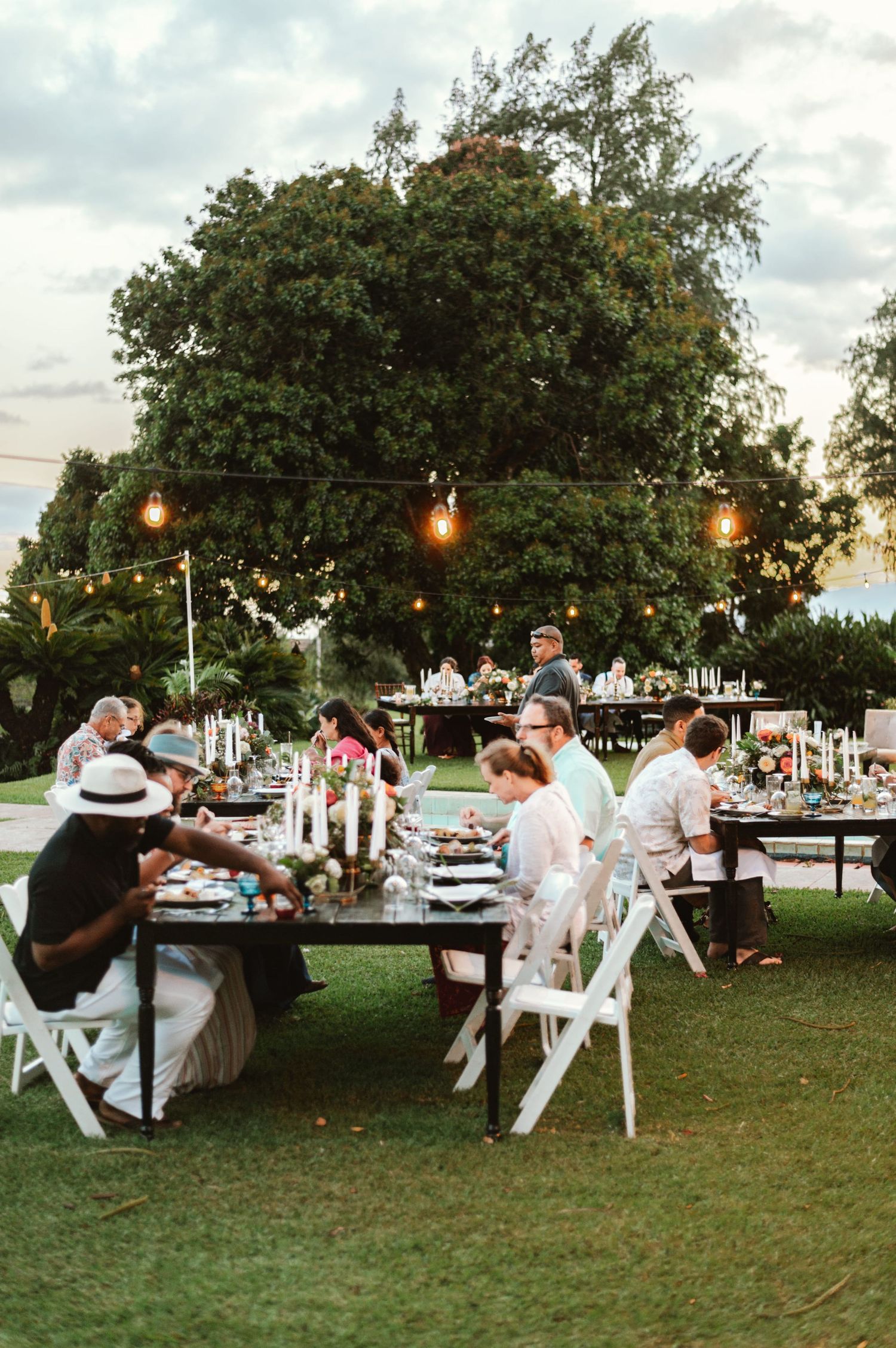 Outdoor garden wedding reception with guests dining at long wooden tables under string lights and a large tree.