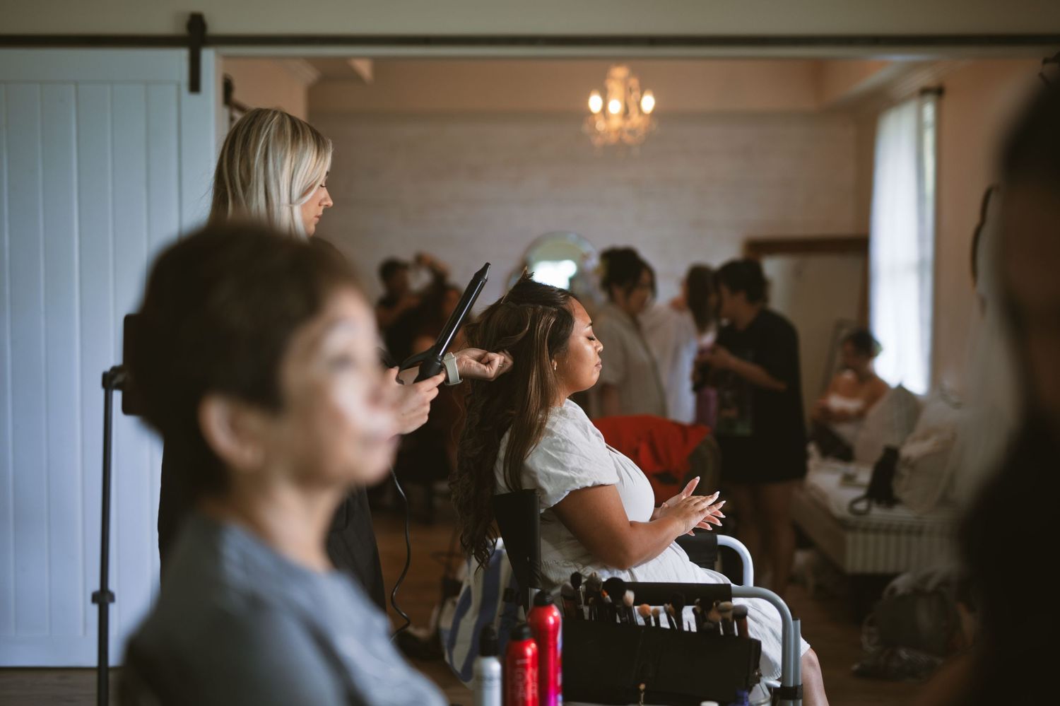 A hairstylist curls a seated woman's hair in a bright wedding preparation room with guests in the background.