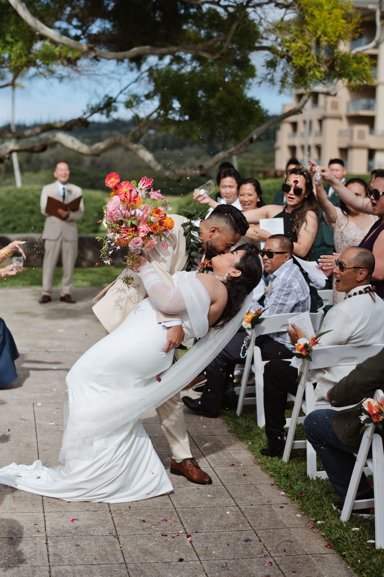 Couple shares romantic dip kiss at outdoor wedding ceremony while guests cheer and toss confetti.