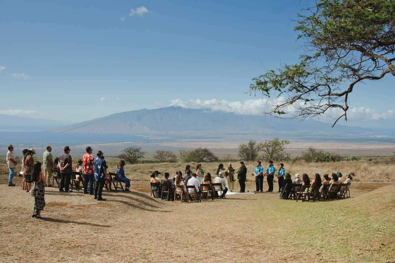 Group of people gathered outdoors in an open savanna landscape with mountain views under a clear blue sky.