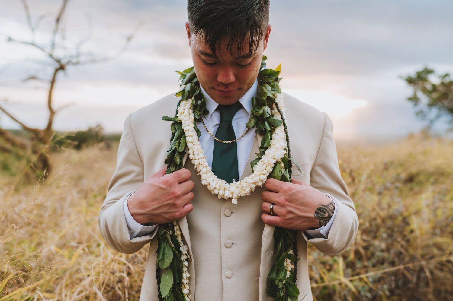 Groom in beige suit wearing traditional Hawaiian lei of white flowers and green leaves outdoors at sunset.