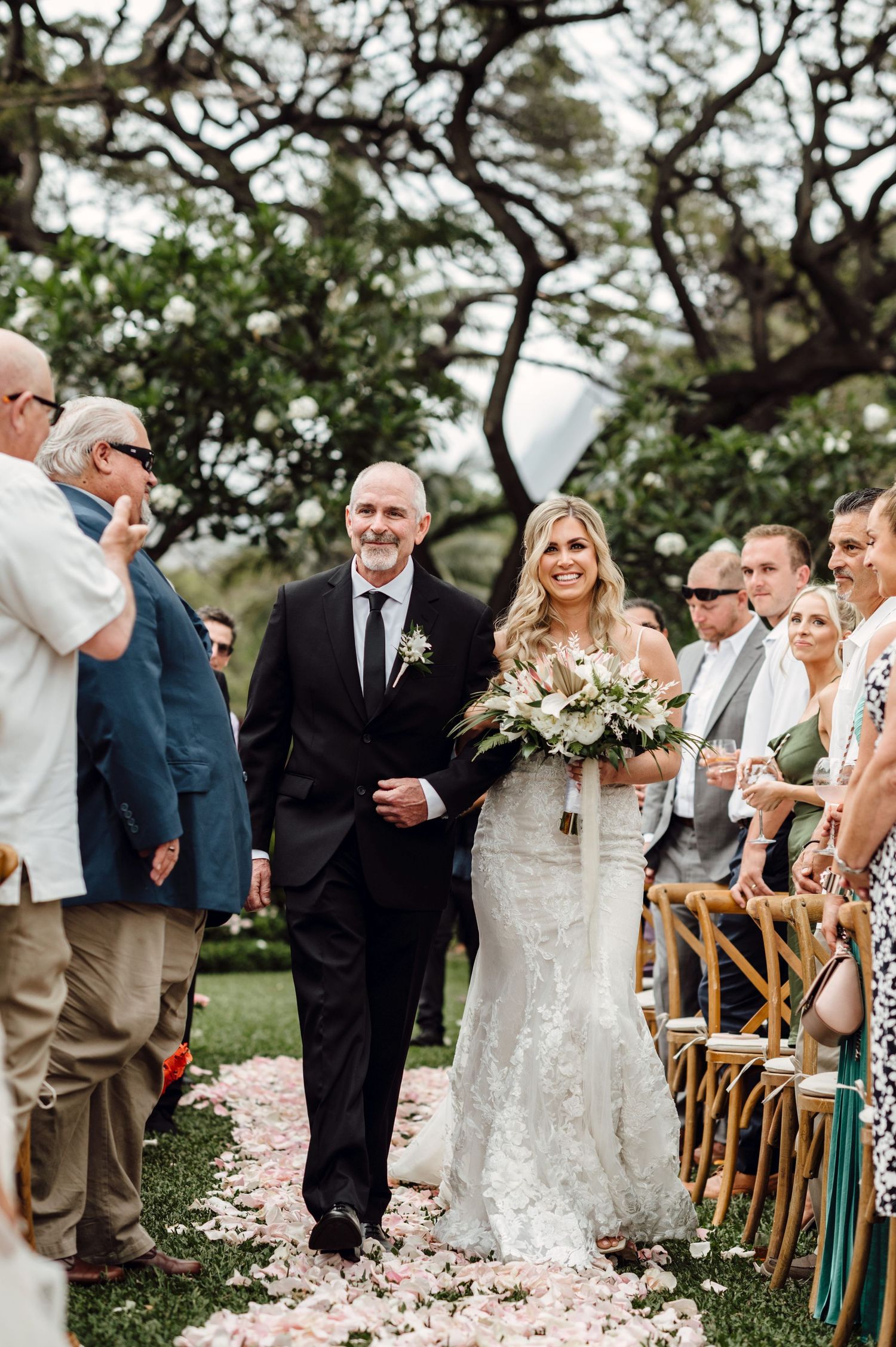 Bride walks down petal-lined aisle with escort at outdoor wedding ceremony amid lush greenery.