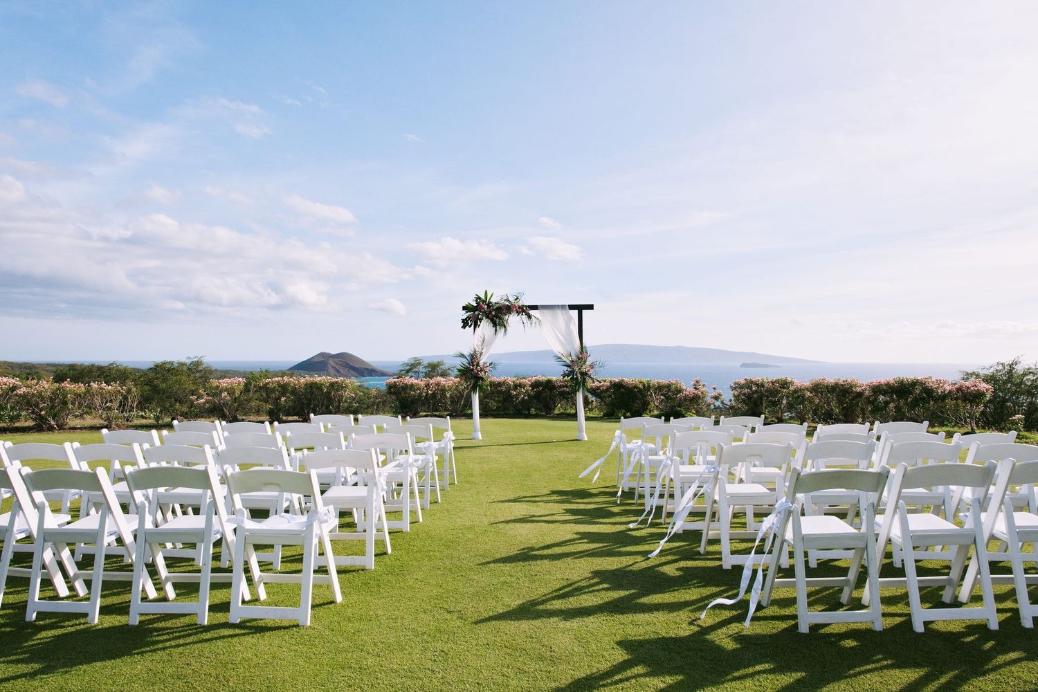 Outdoor wedding ceremony setup with white folding chairs on green lawn, floral arch, and ocean view under blue sky.