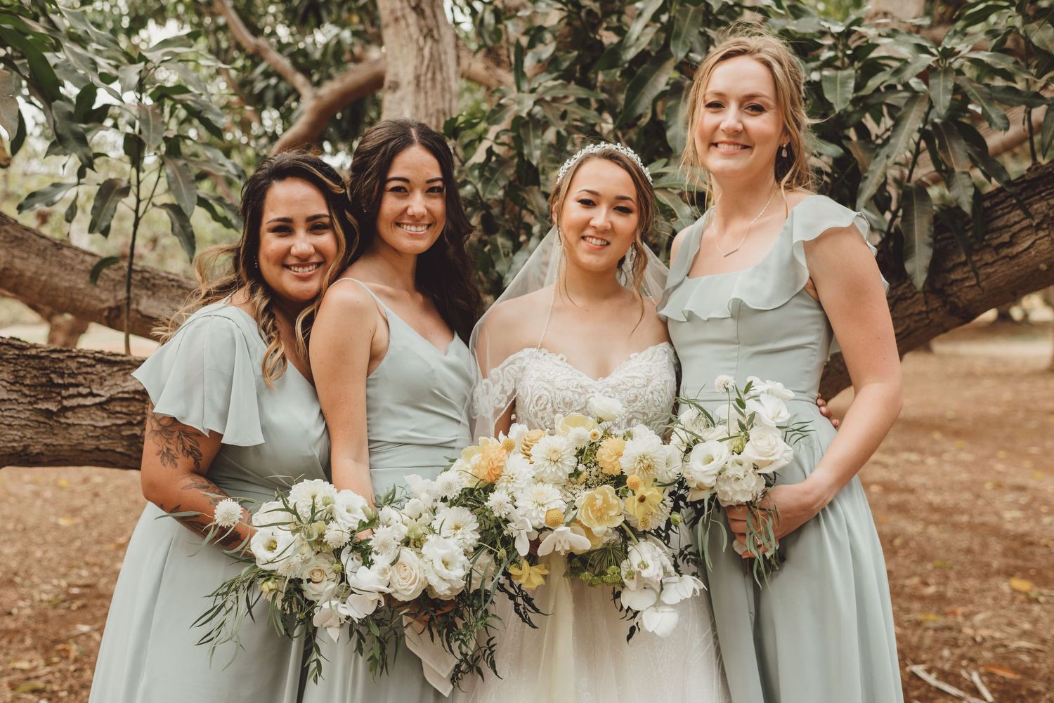 Bride in white gown with three bridesmaids in sage green dresses holding white and yellow bouquets outdoors.