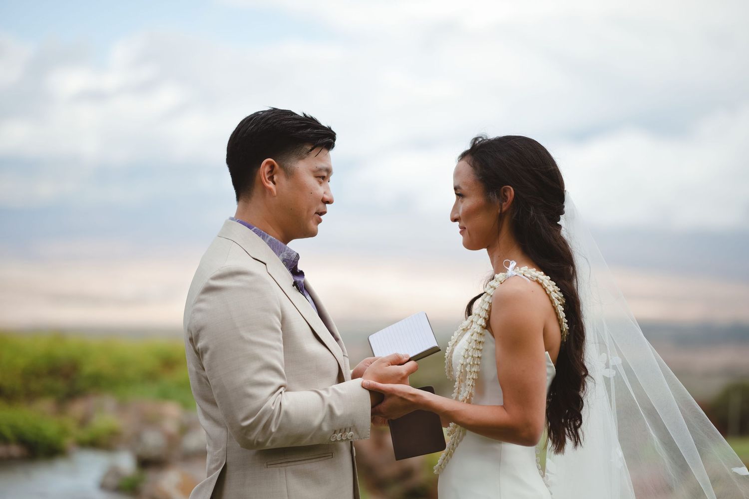 Asian couple exchanging vows outdoors, groom in tan suit, bride in white wedding dress with veil, scenic landscape backdrop.