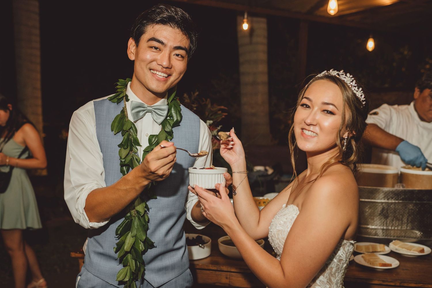 Bride and groom smiling while sharing dessert at their wedding reception, groom wearing lei and blue vest.