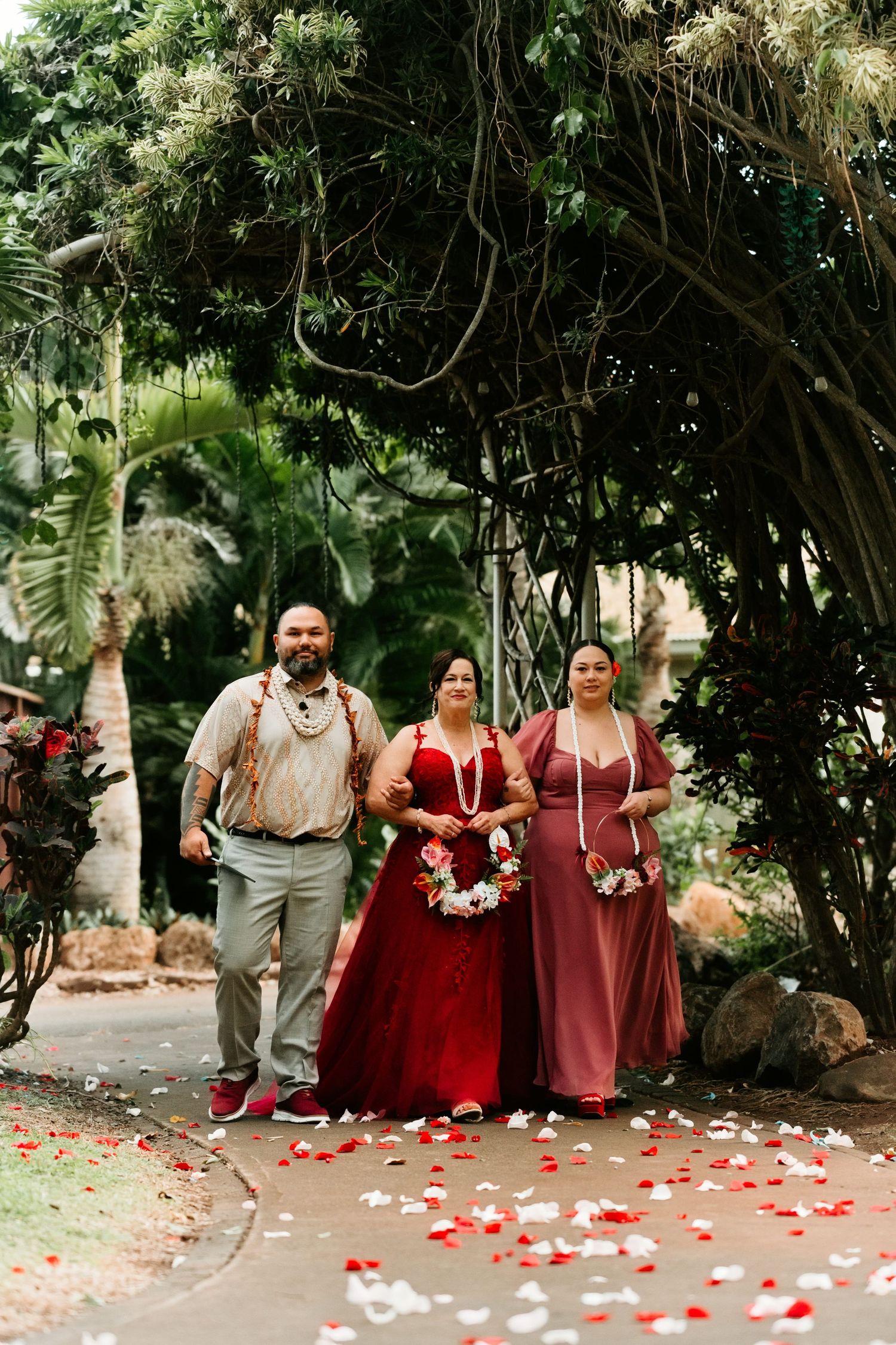 Three people in formal attire walk down a petal-lined path under lush tropical trees at an outdoor Hawaiian wedding.