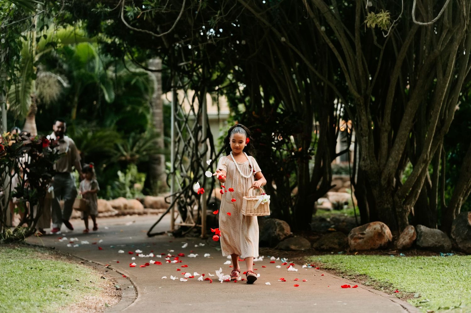 A flower girl in a white dress scatters red rose petals on a garden path lined with lush green trees.