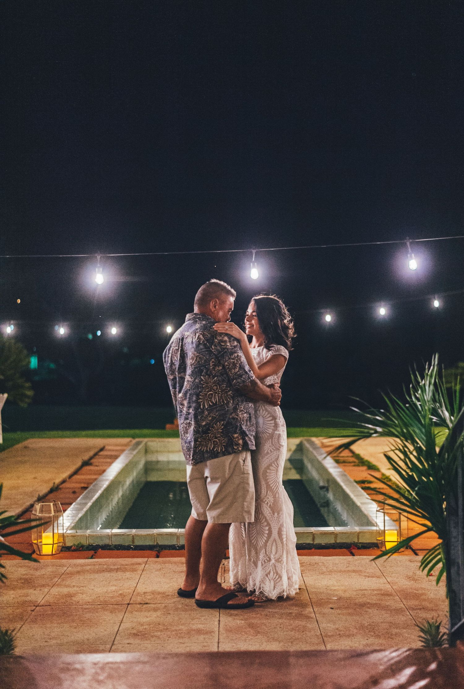 Intimate nighttime dance moment between couple under string lights by a pool.