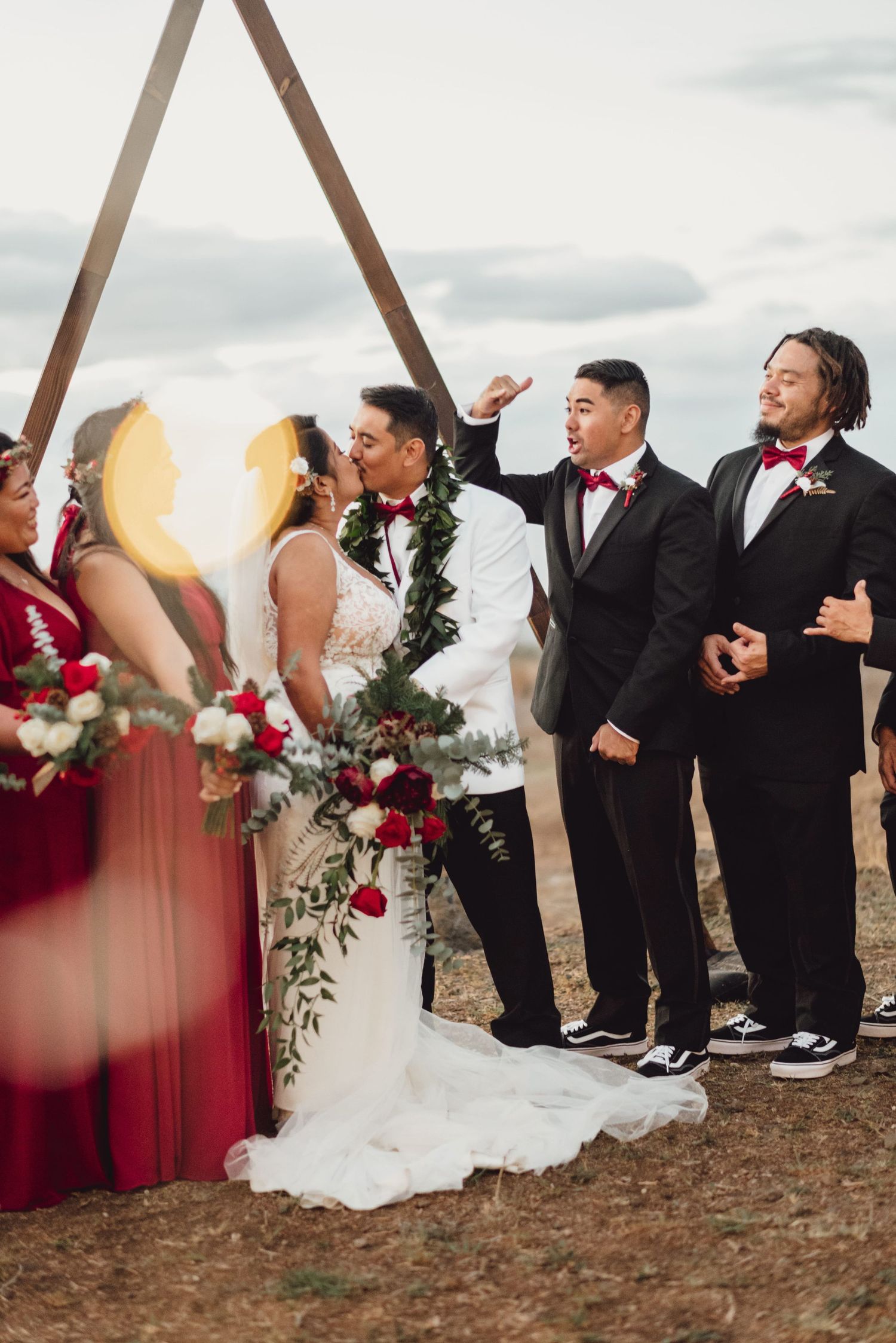 Wedding party celebrates as bride and groom share first kiss under a triangular arch with red and white floral decor.