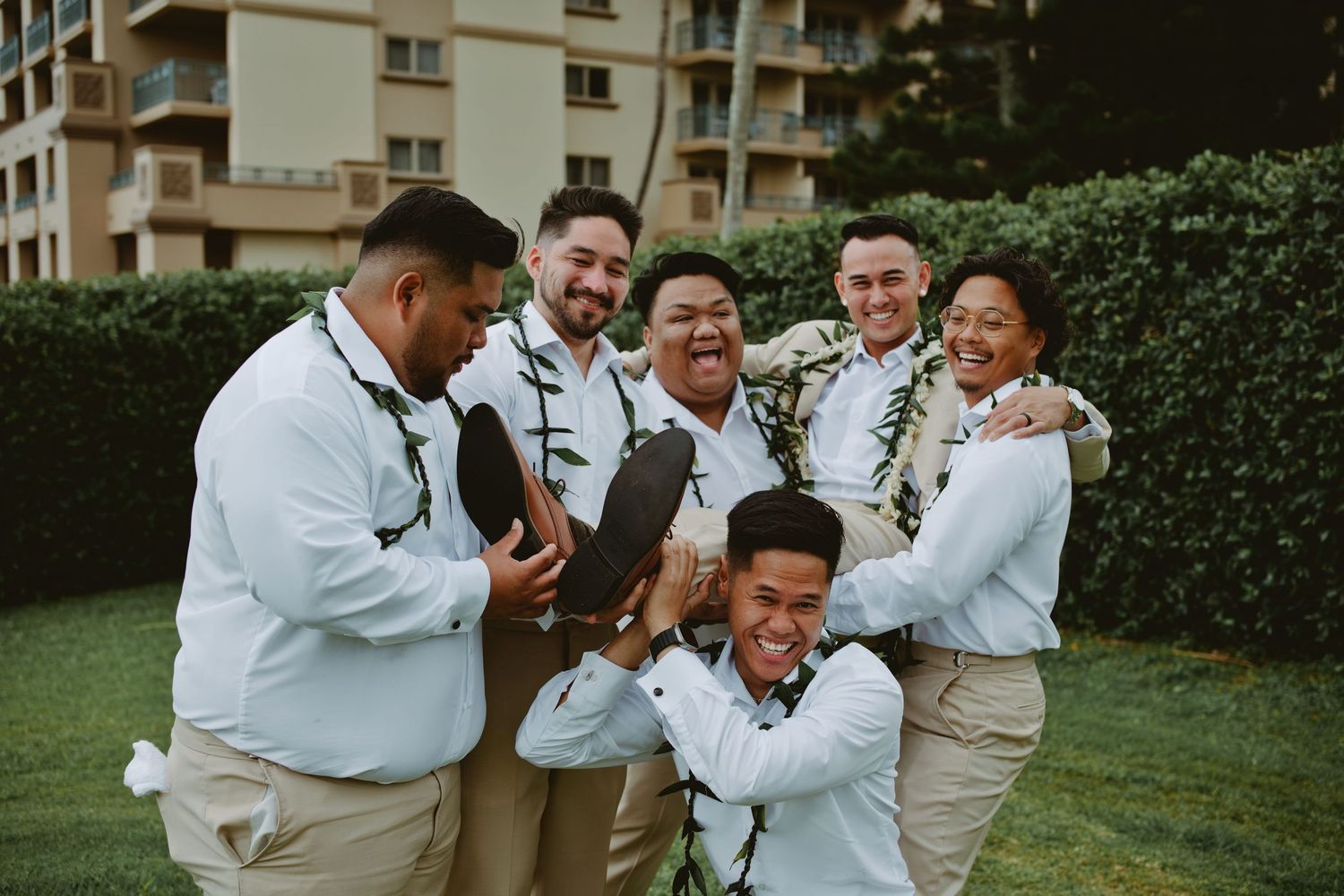 Groomsmen in white shirts and khaki pants laughing outdoors at a Hawaiian wedding venue.