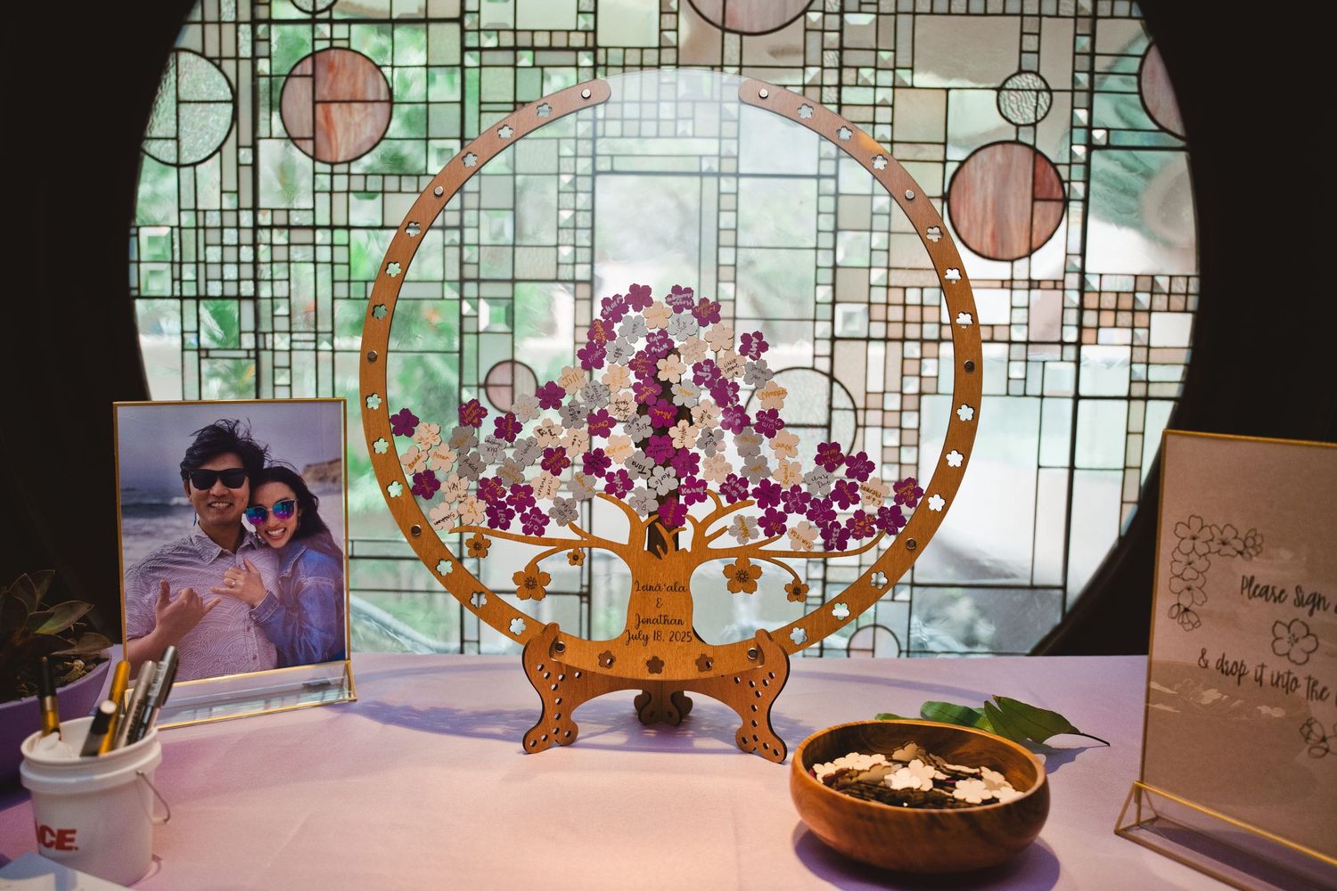 Decorative wooden wedding guest book tree with purple flowers in a circular frame, displayed on a table with a couple photo.