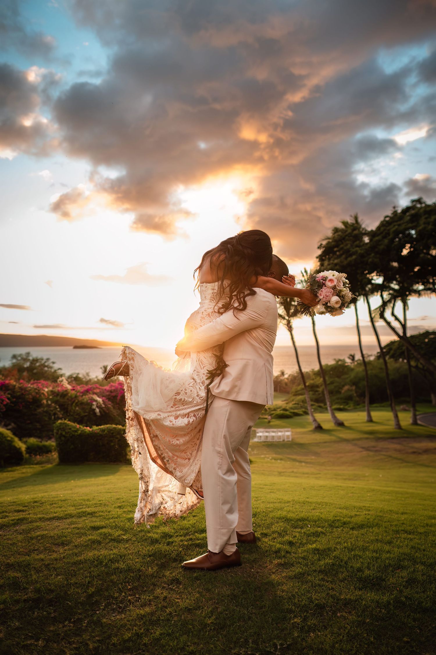 Couple sharing a romantic embrace at sunset on a lush green lawn, bride in lace gown holding bouquet, groom in ivory suit.
