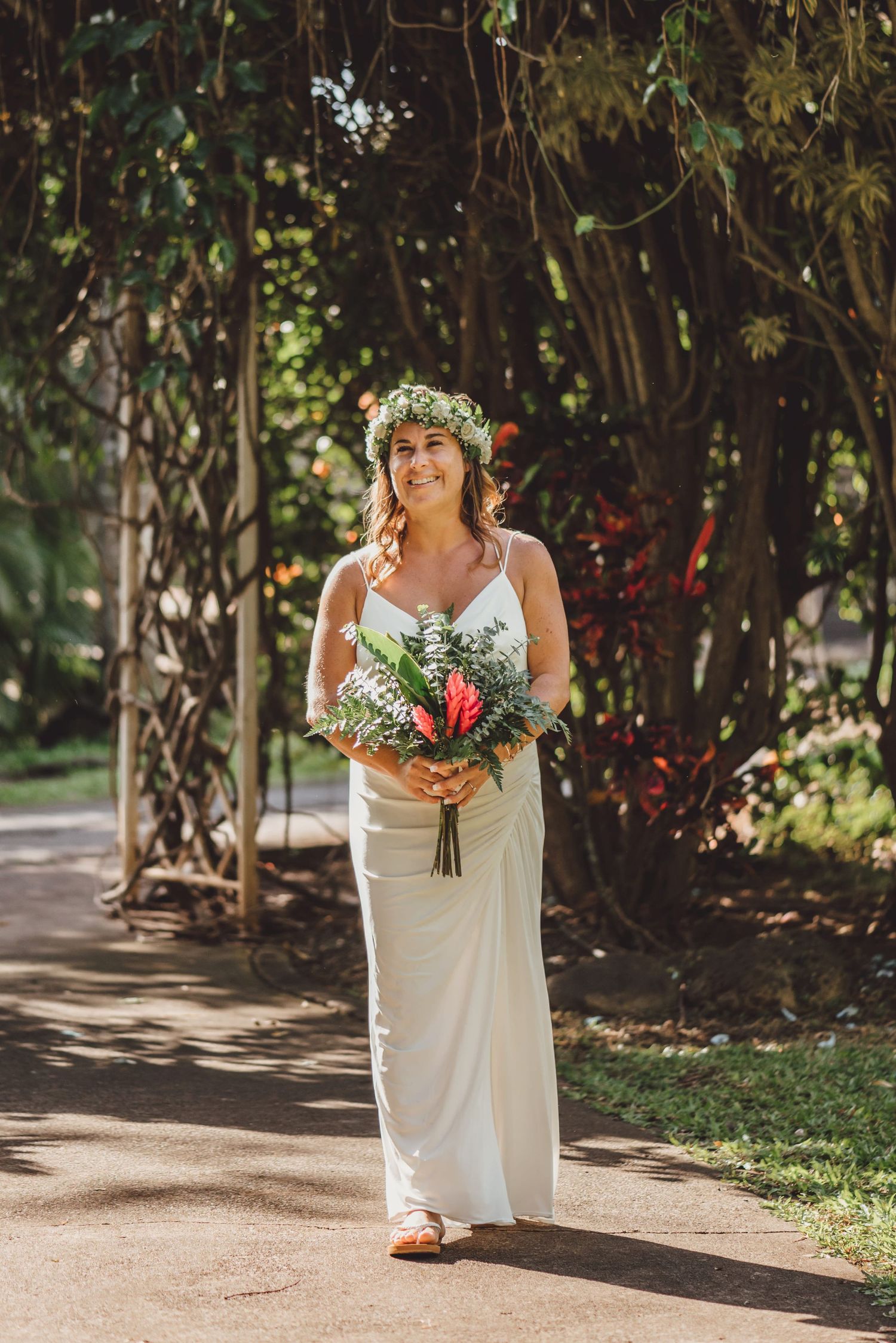 Bride in white gown and floral crown holds tropical bouquet on sunlit garden path.
