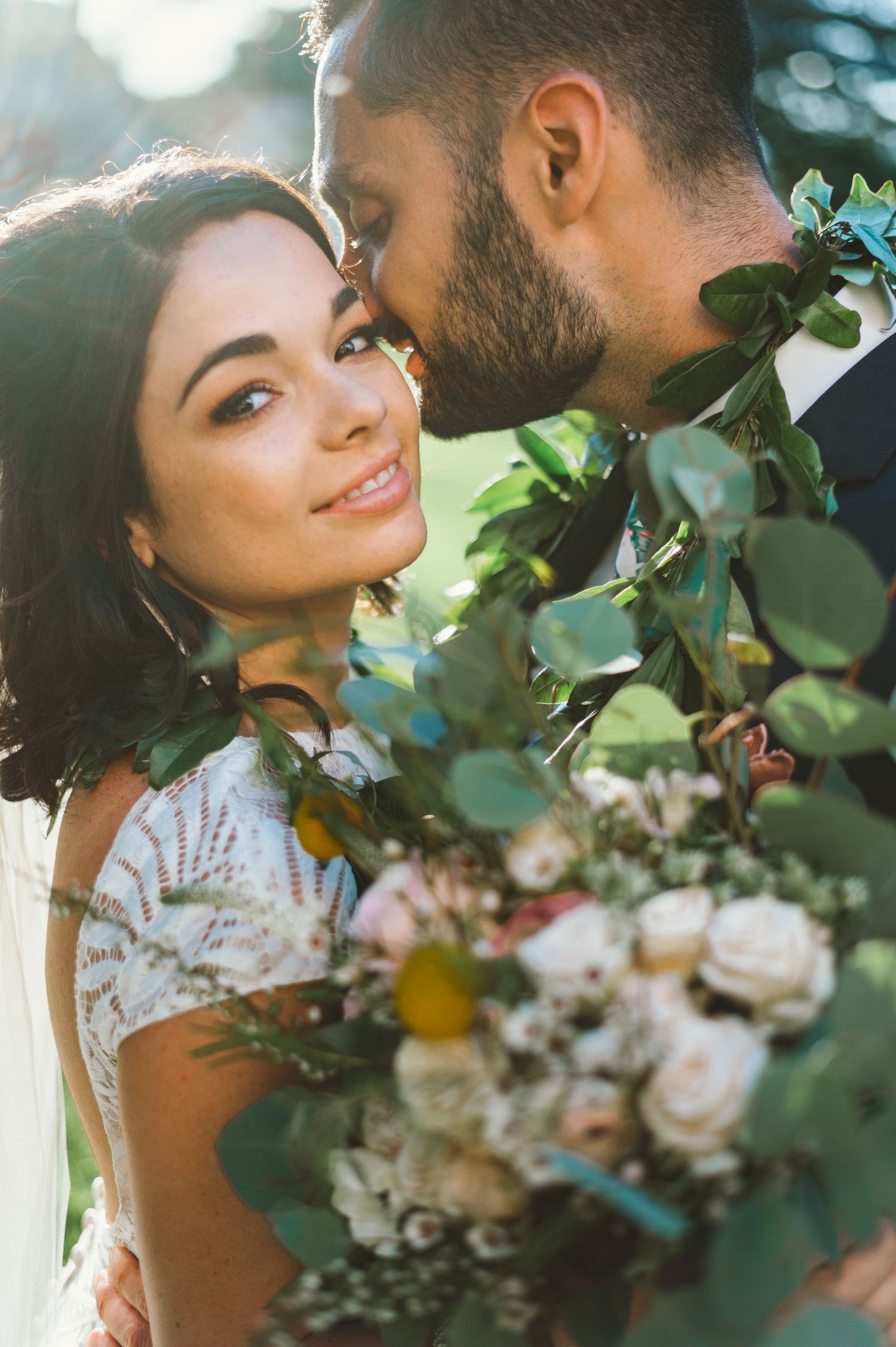 A bride smiles softly as her groom kisses her cheek, surrounded by lush greenery and a romantic bouquet.