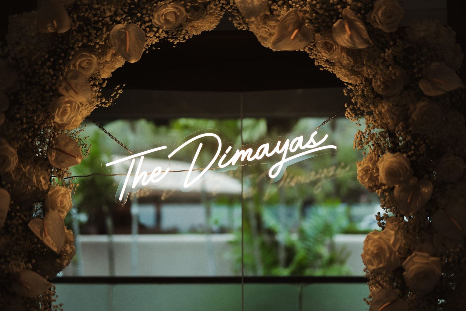 Elegant neon sign reading The Dimayas framed by a floral arch with white roses and greenery at a wedding reception.