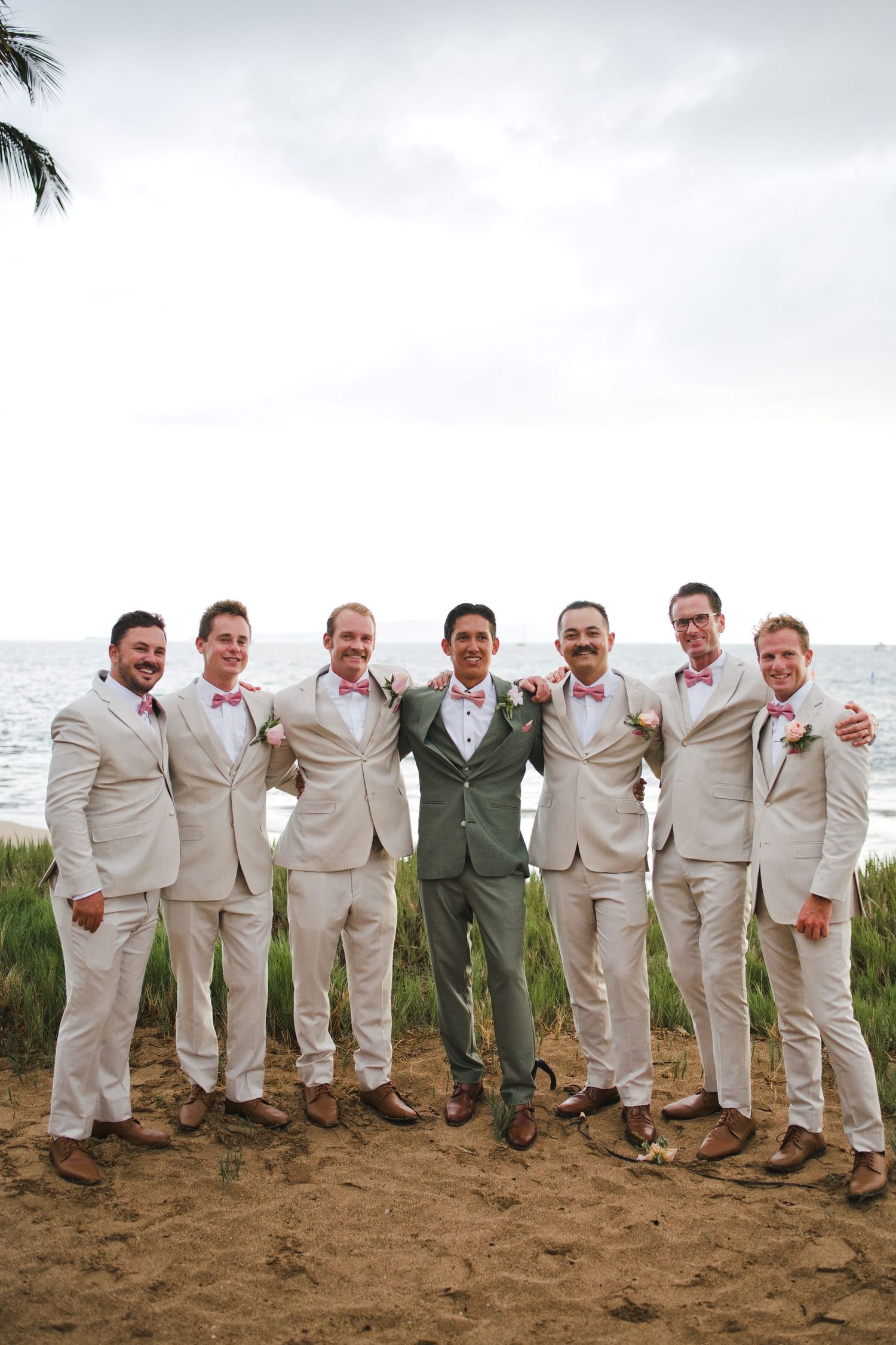 Groom in green suit poses with six groomsmen in cream suits and pink bow ties by the ocean.