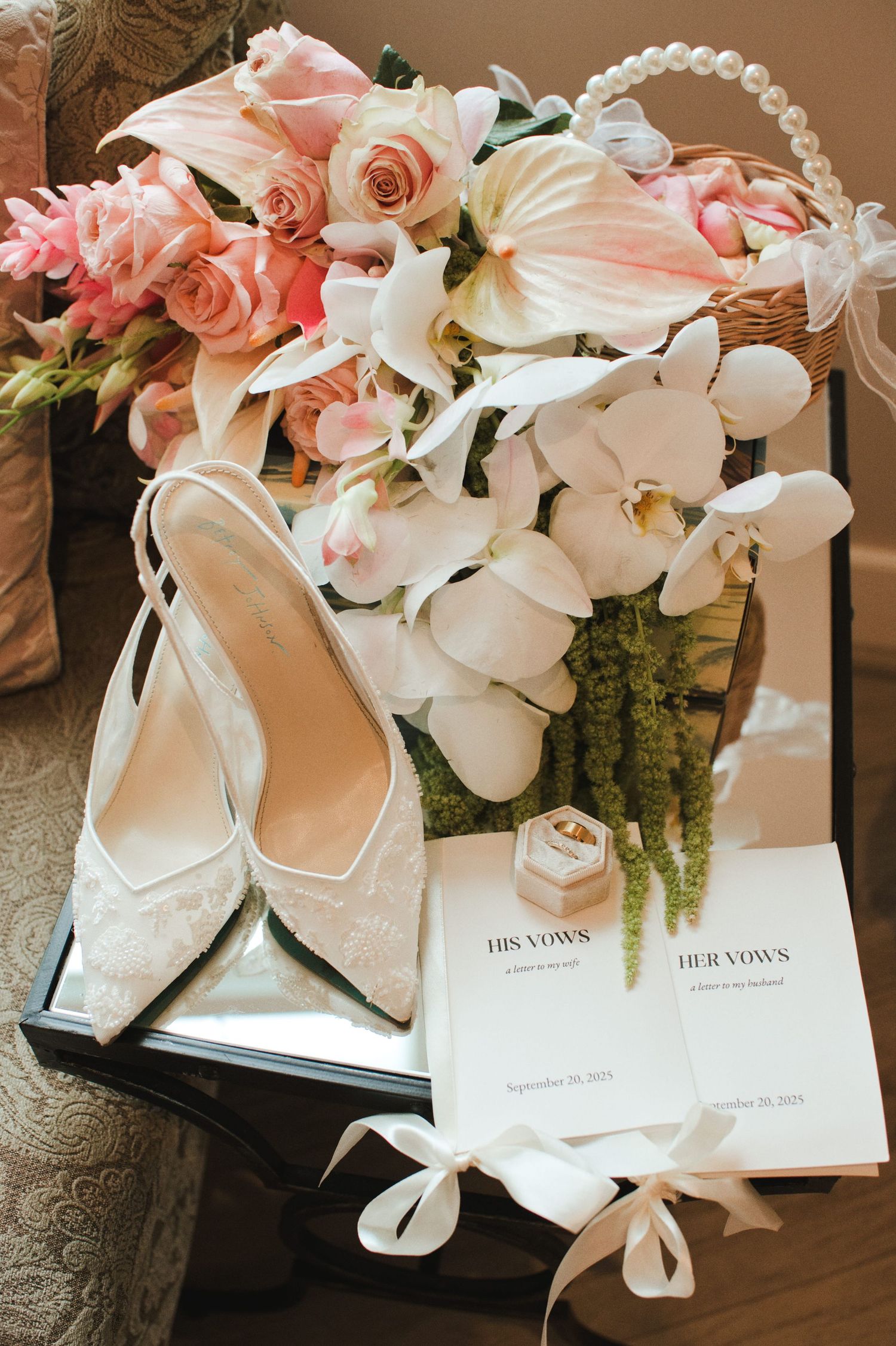 Wedding flat lay with lace bridal heels, vow books, rings, and pink floral bouquet arrangement.