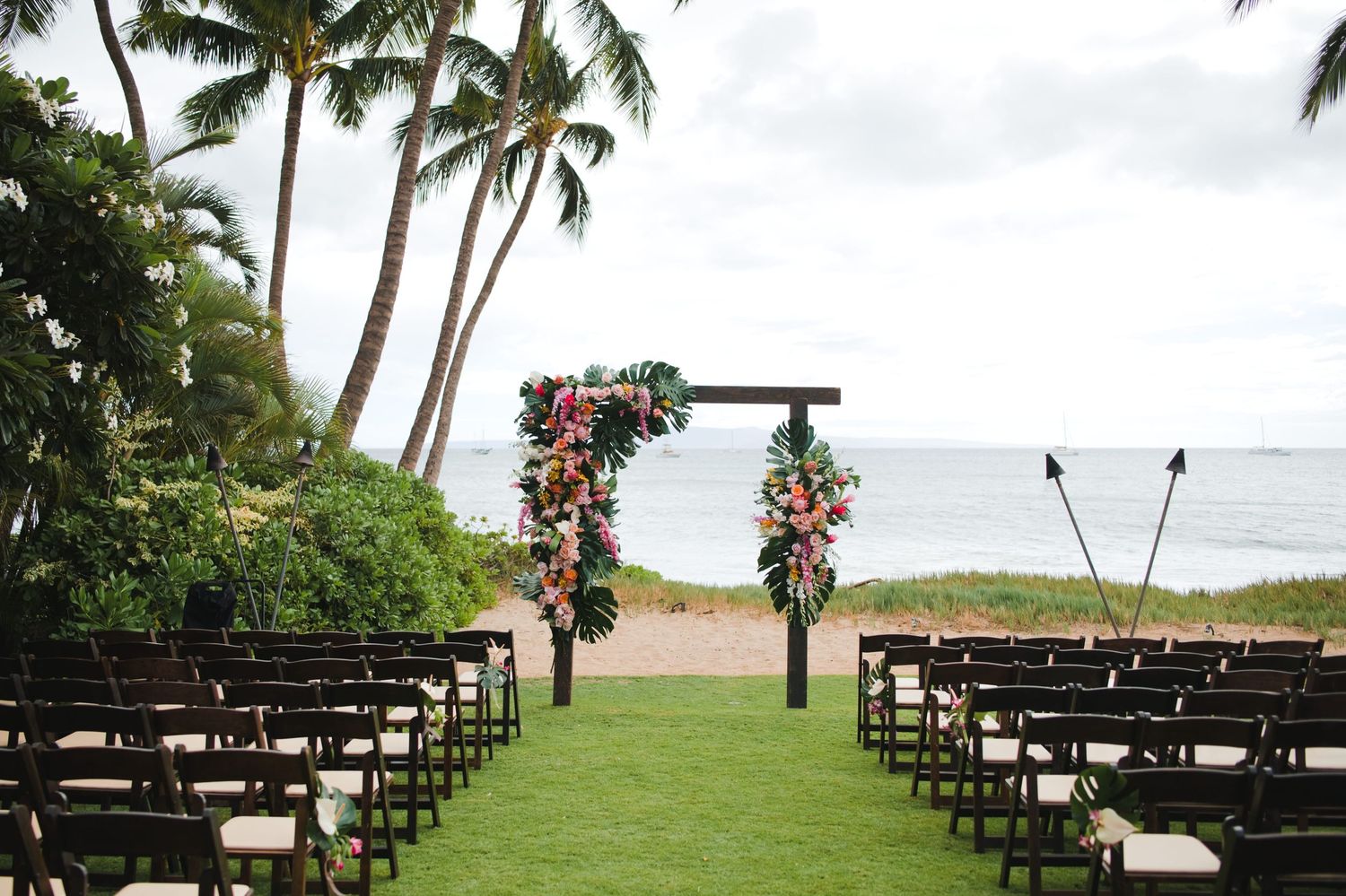 Outdoor beach wedding ceremony setup with floral arch, dark wood chairs on green lawn, palm trees, and ocean view in Hawaii.