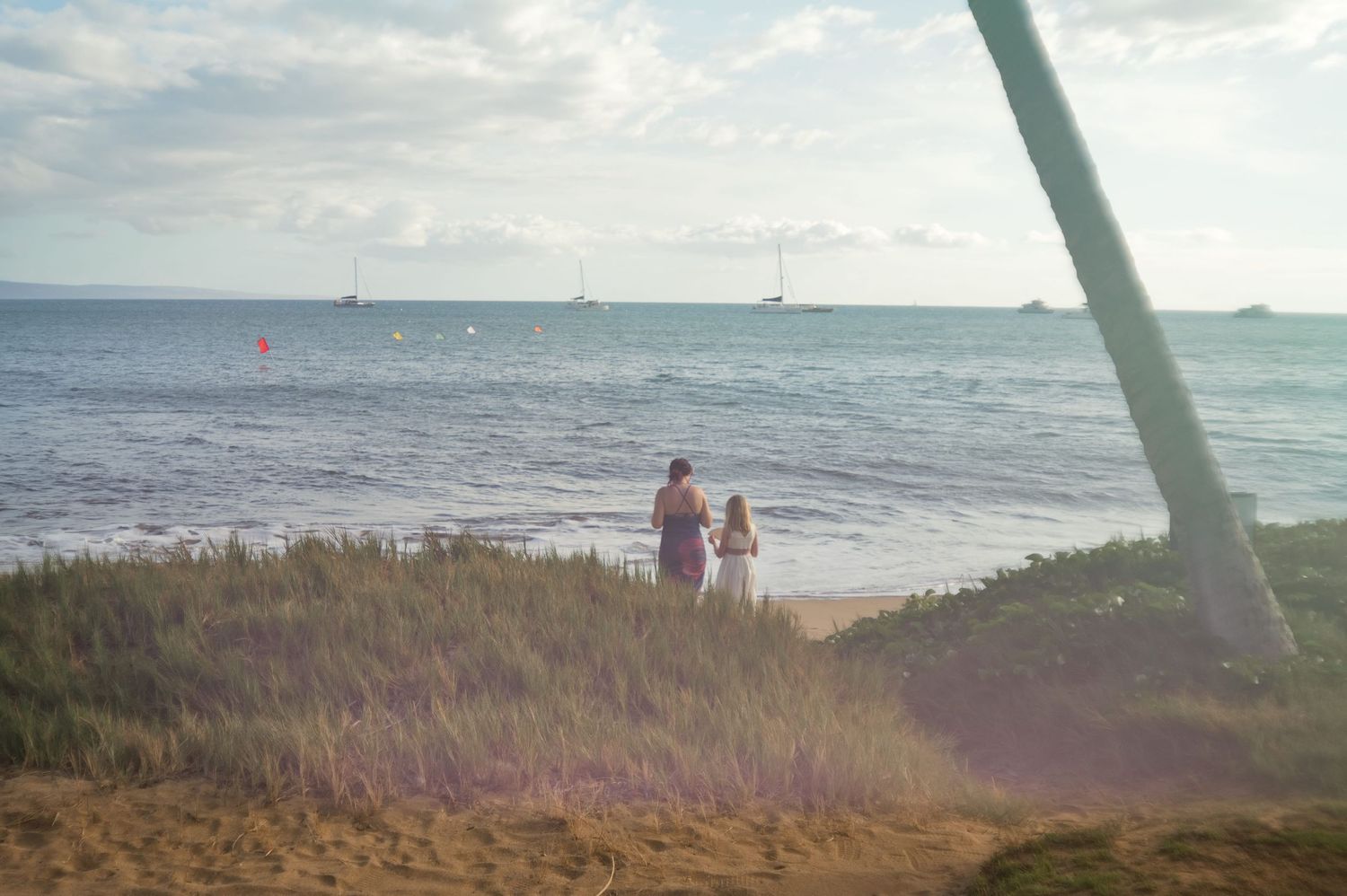 Two people stand on a sandy beach in Hawaii watching sailboats on the ocean under a partly cloudy sky.