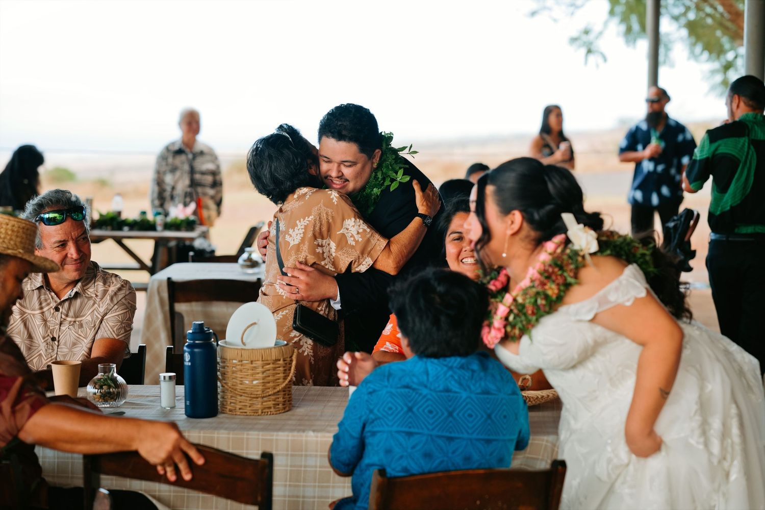 Bride in white gown with lei shares joyful hugs with guests at an outdoor Hawaiian wedding reception.