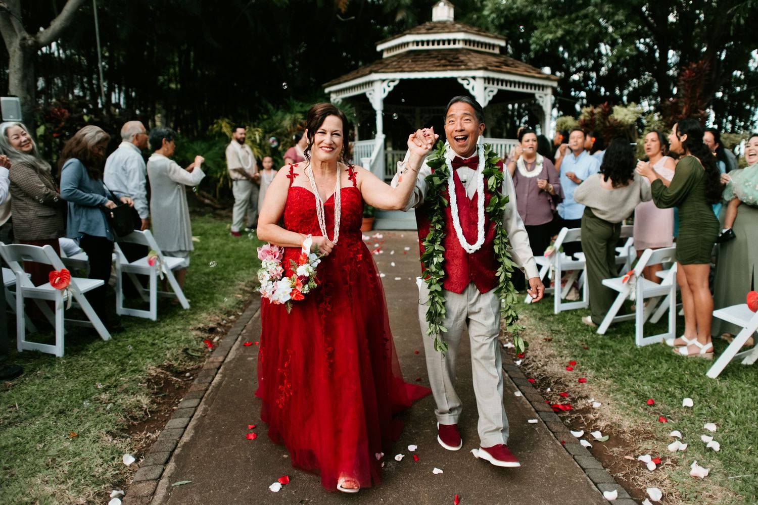Joyful newlyweds walk down outdoor aisle wearing red dress and lei garlands at Hawaiian gazebo wedding ceremony.