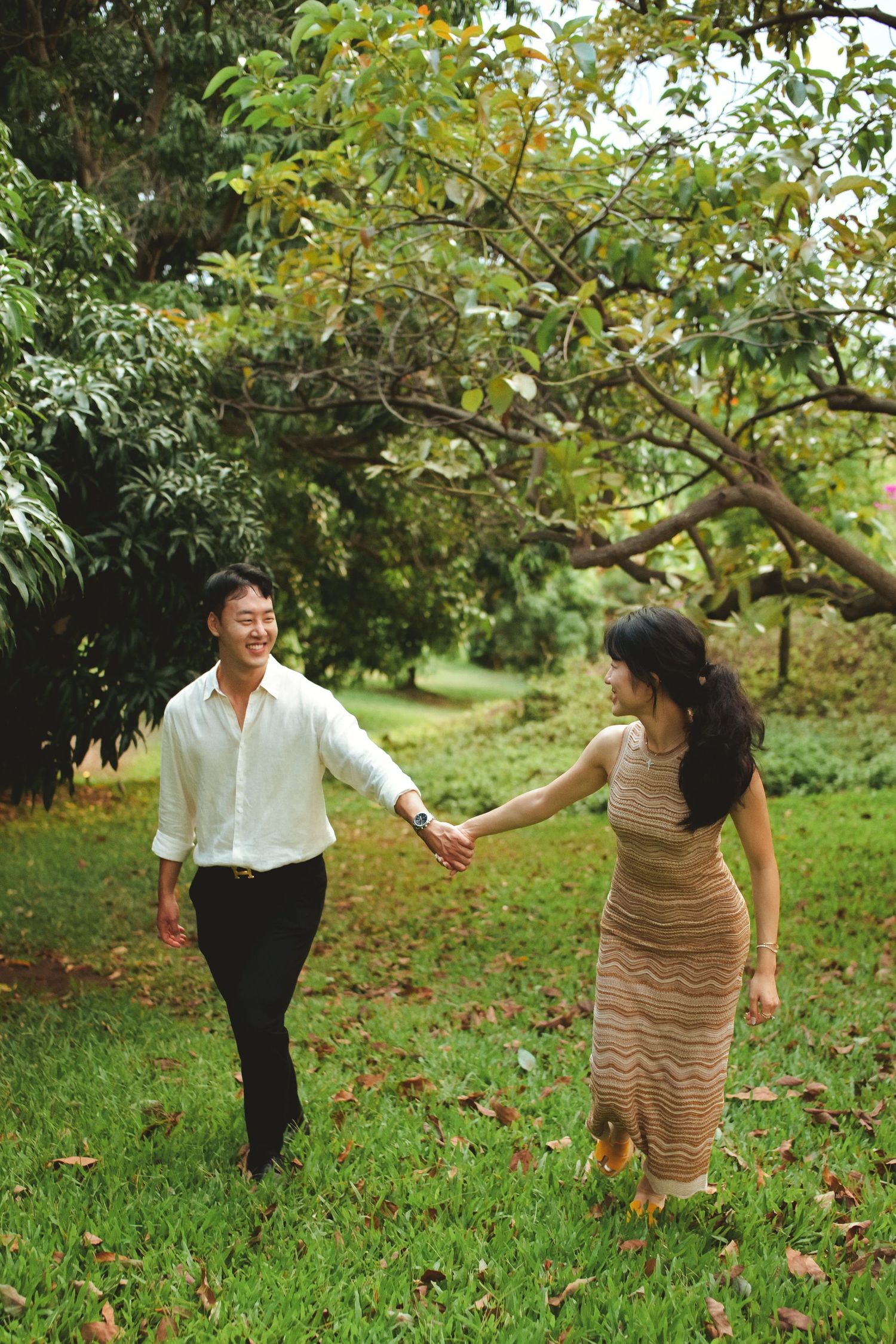 Asian couple holding hands and walking through a lush green park, smiling joyfully on a romantic outdoor stroll.