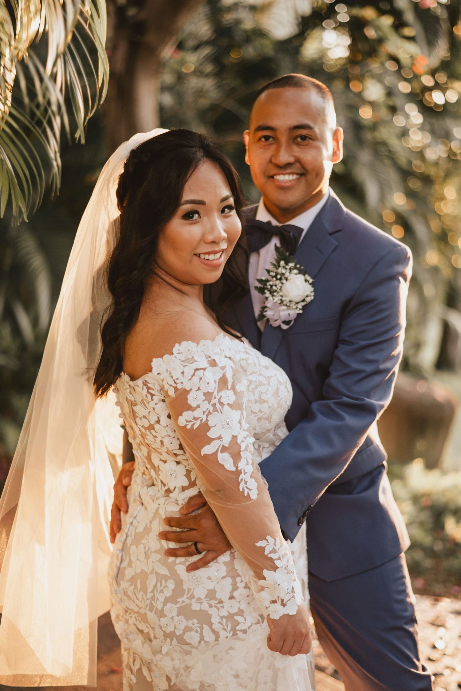 Bride in lace gown and veil poses with groom in blue suit and boutonniere in a sunlit garden setting.