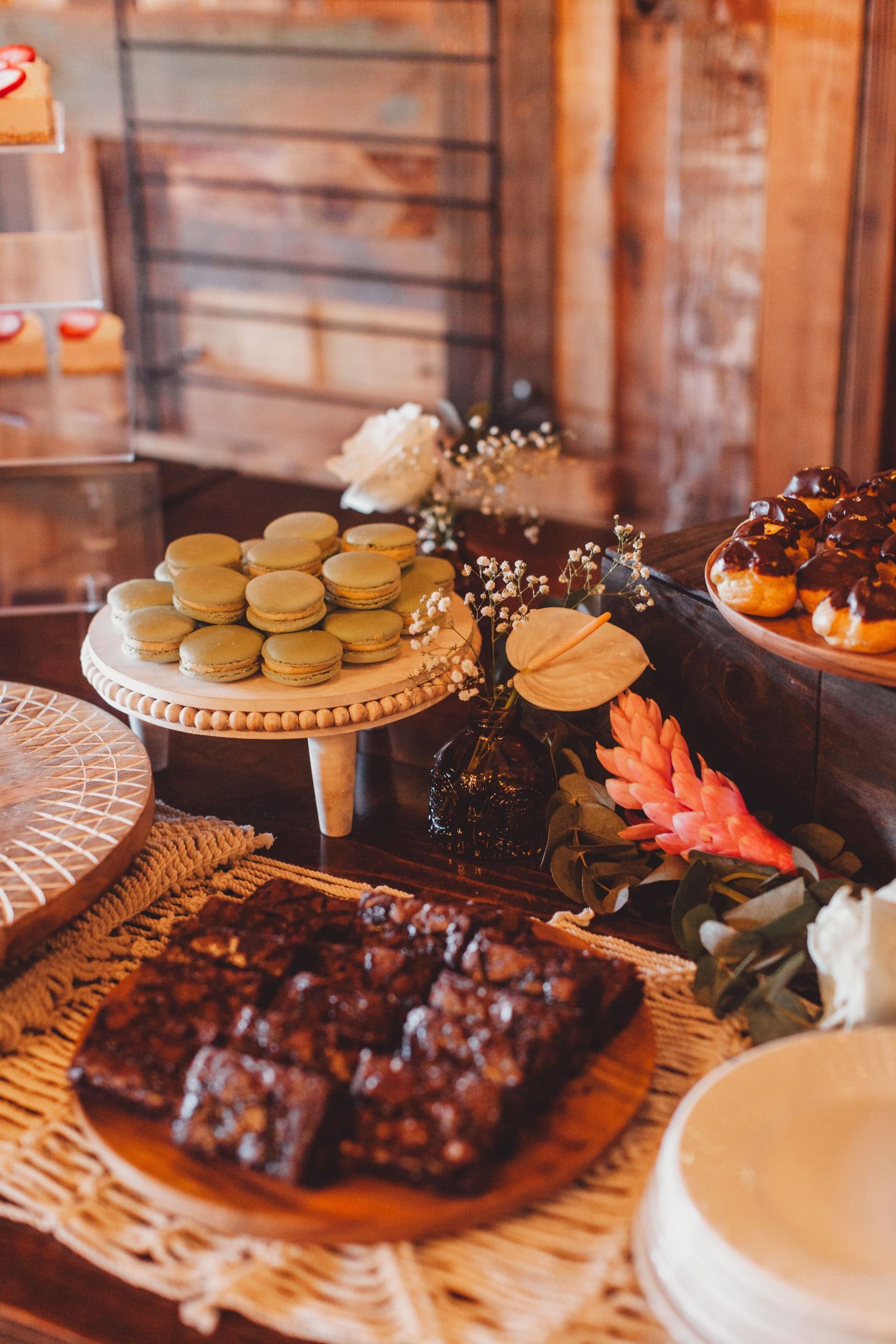 Rustic dessert table with macarons, chocolate brownies, and glazed donuts displayed on wooden stands with floral decor.