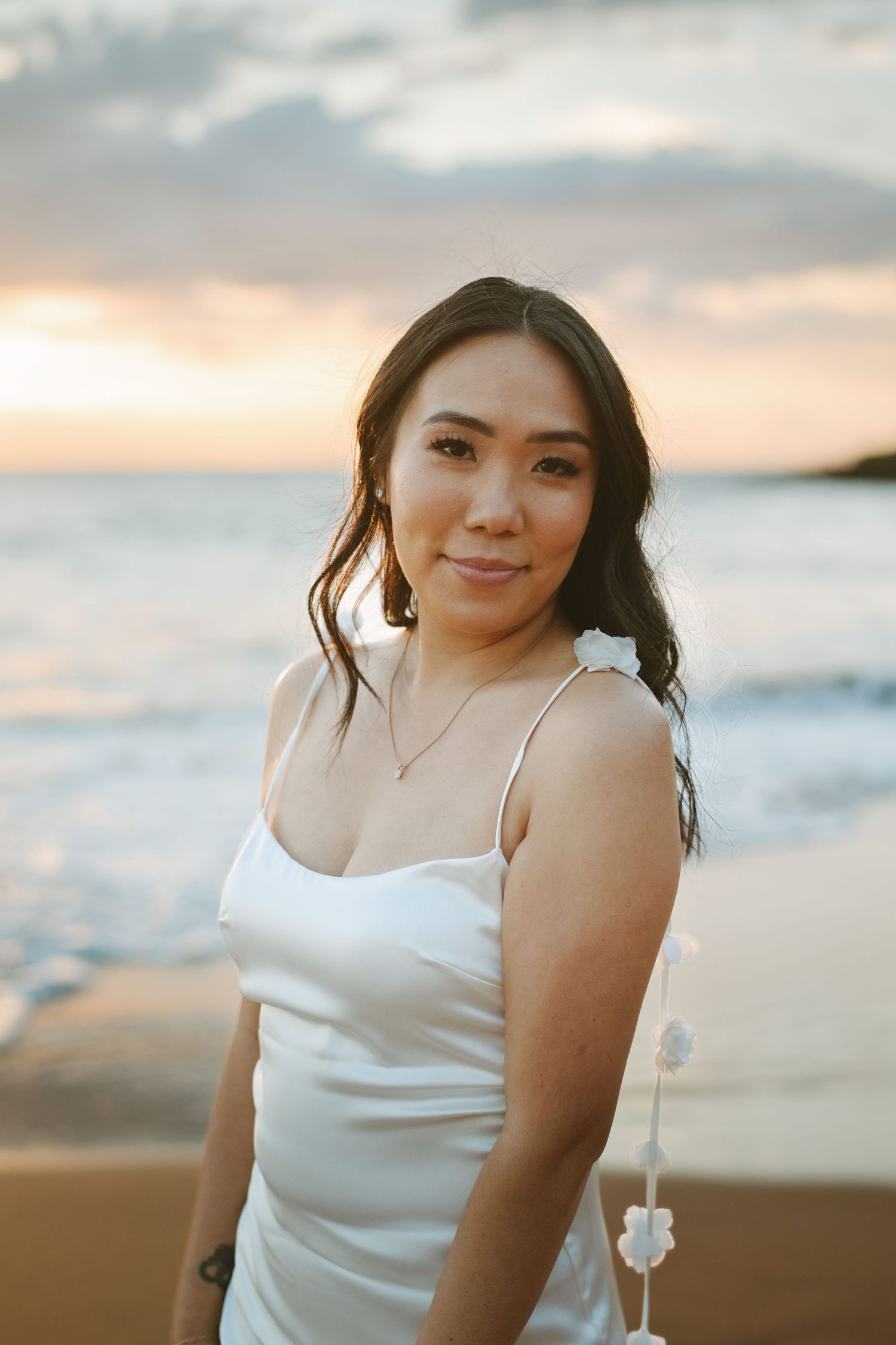 Woman in white satin dress holding flowers on beach during a colorful sunset.