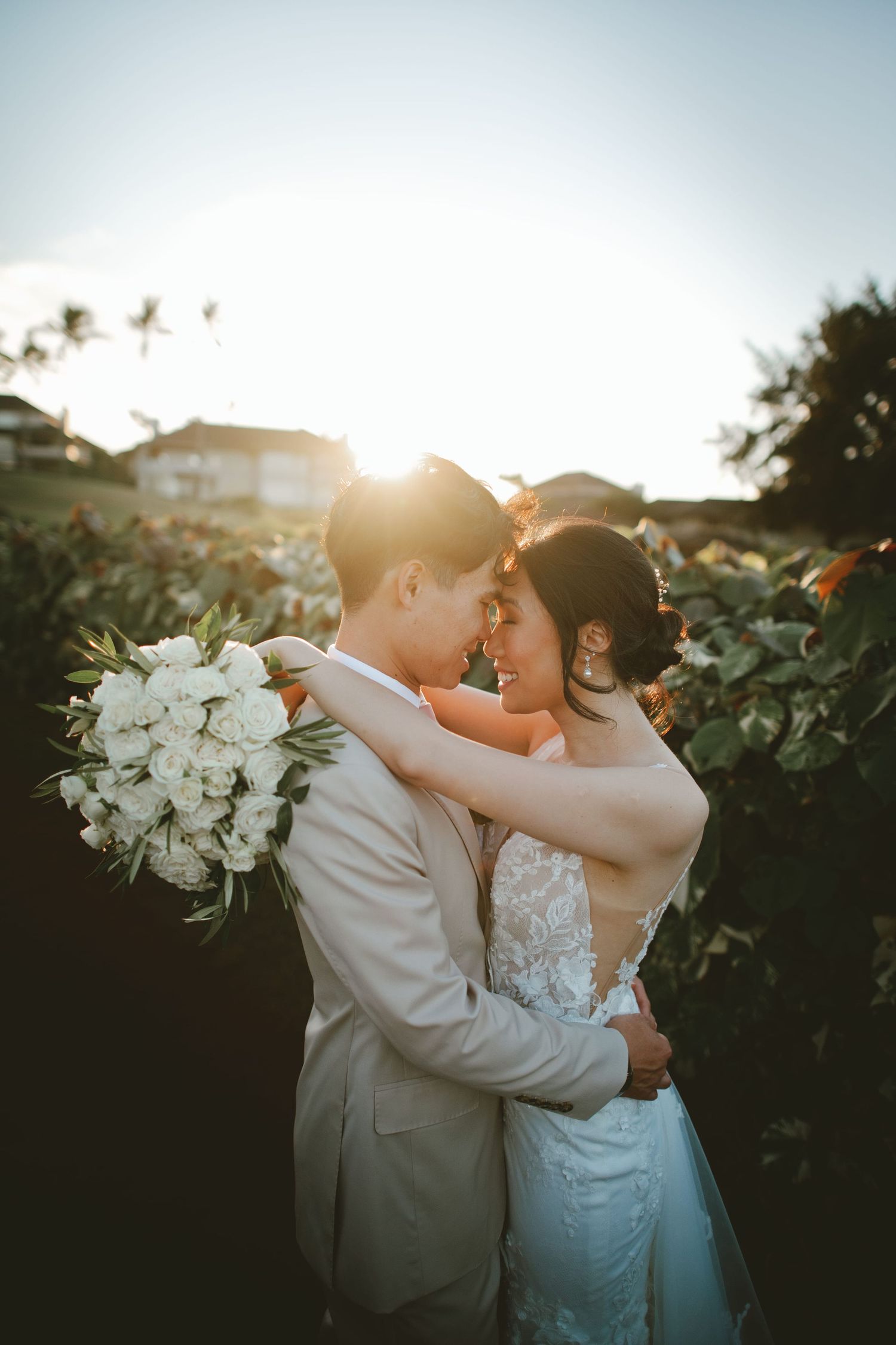 Bride and groom embrace at golden hour sunset, she holds white rose bouquet in lush garden setting.